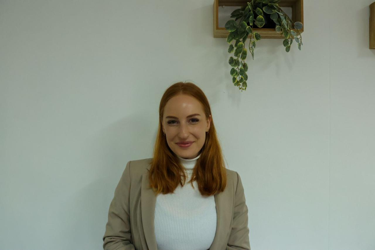 Young woman with shoulder-length red hair smiling, wearing a white turtleneck and beige blazer, standing against a plain wall with hanging plants.