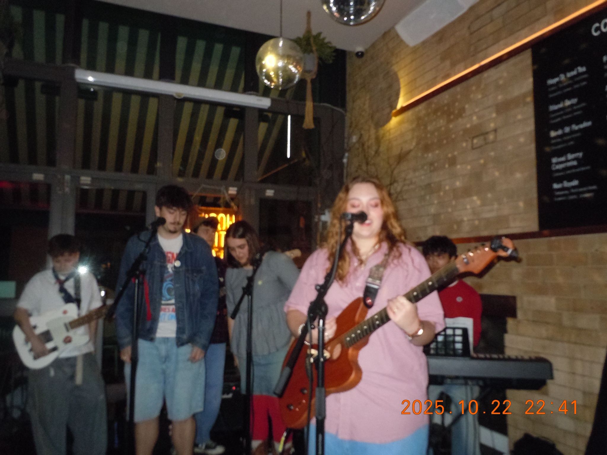 Group of young people performing music on stage in a dimly lit venue with a brick wall and a menu board.