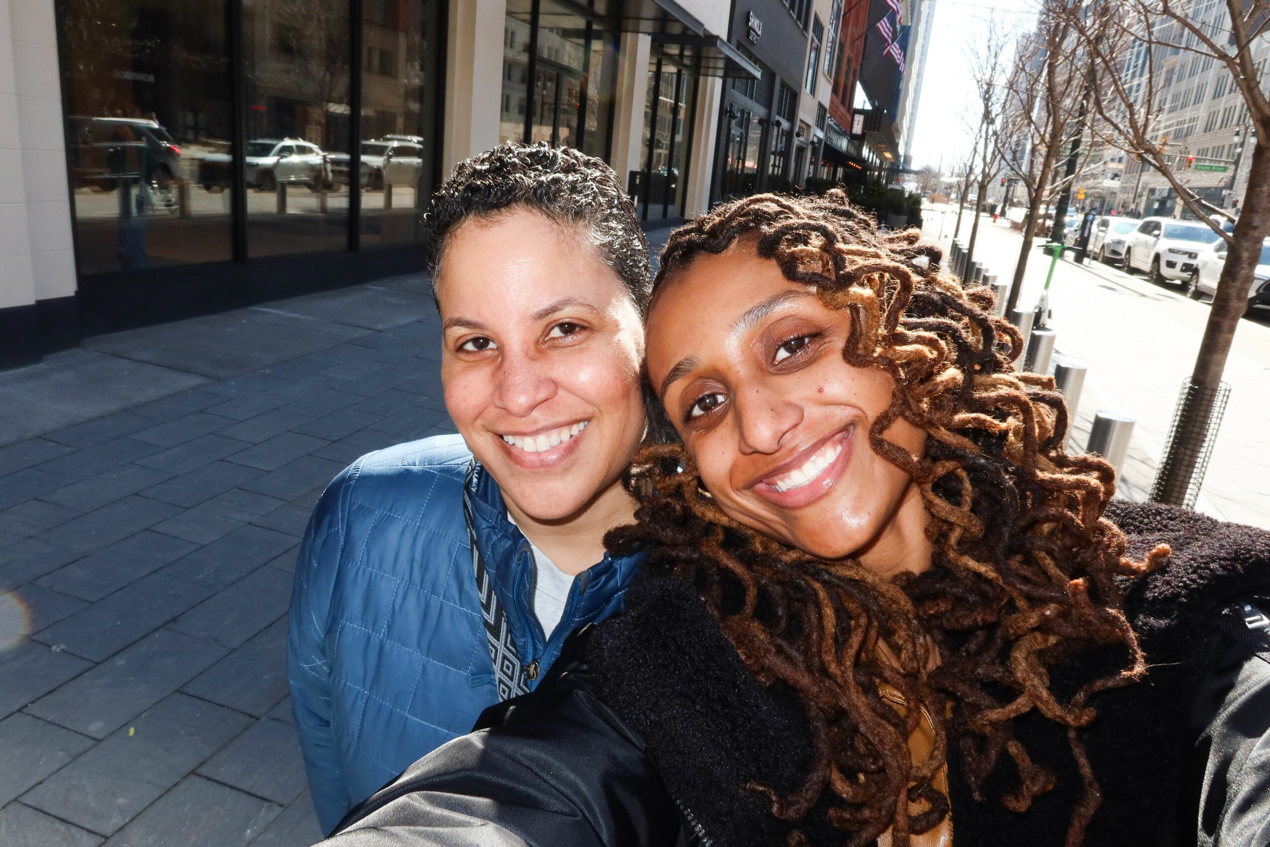 Two women smiling outdoors on a city sidewalk, with buildings and trees in the background.