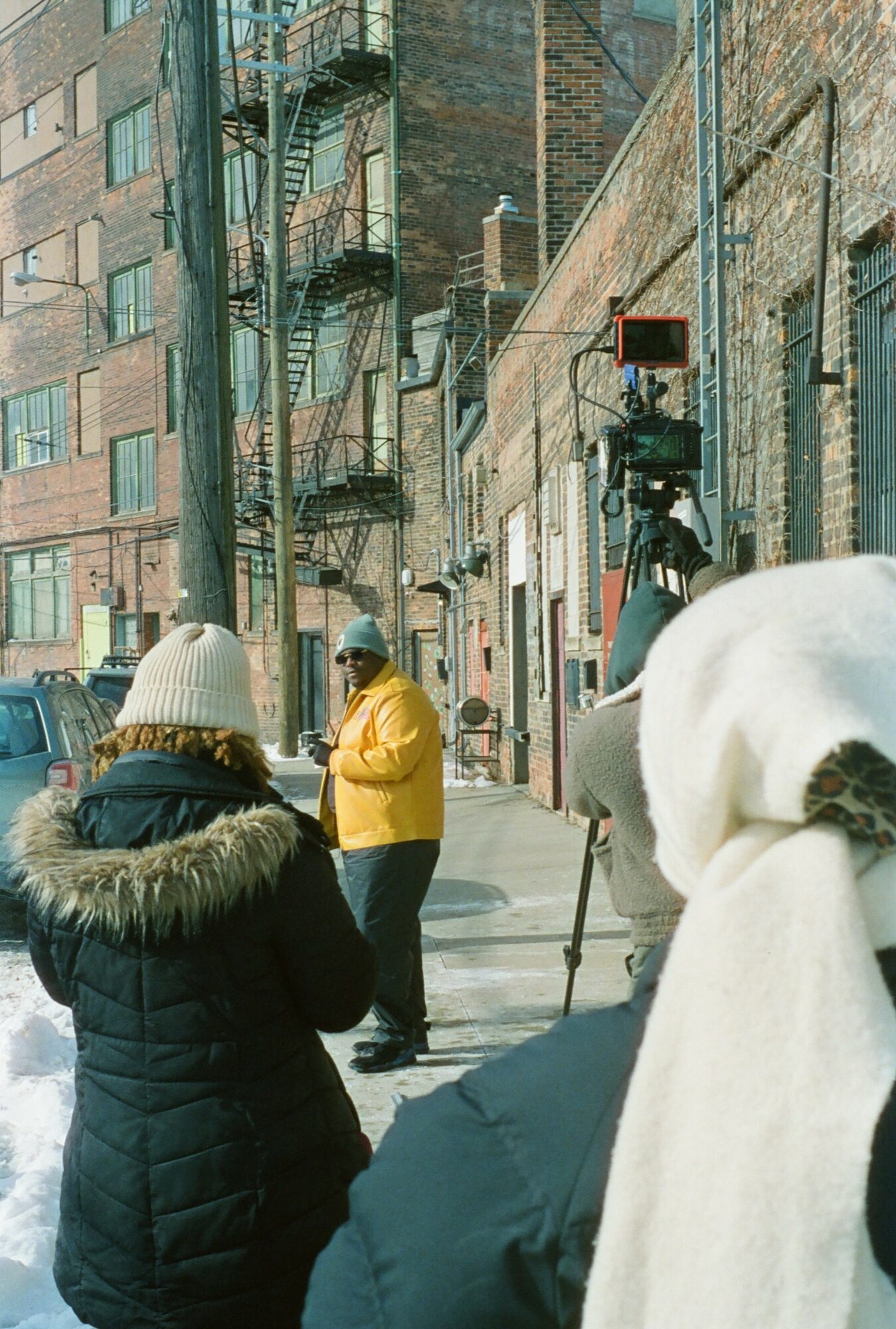 Two people in winter clothing stand on a city sidewalk, with a person in a yellow jacket speaking, and a camera on a tripod nearby.