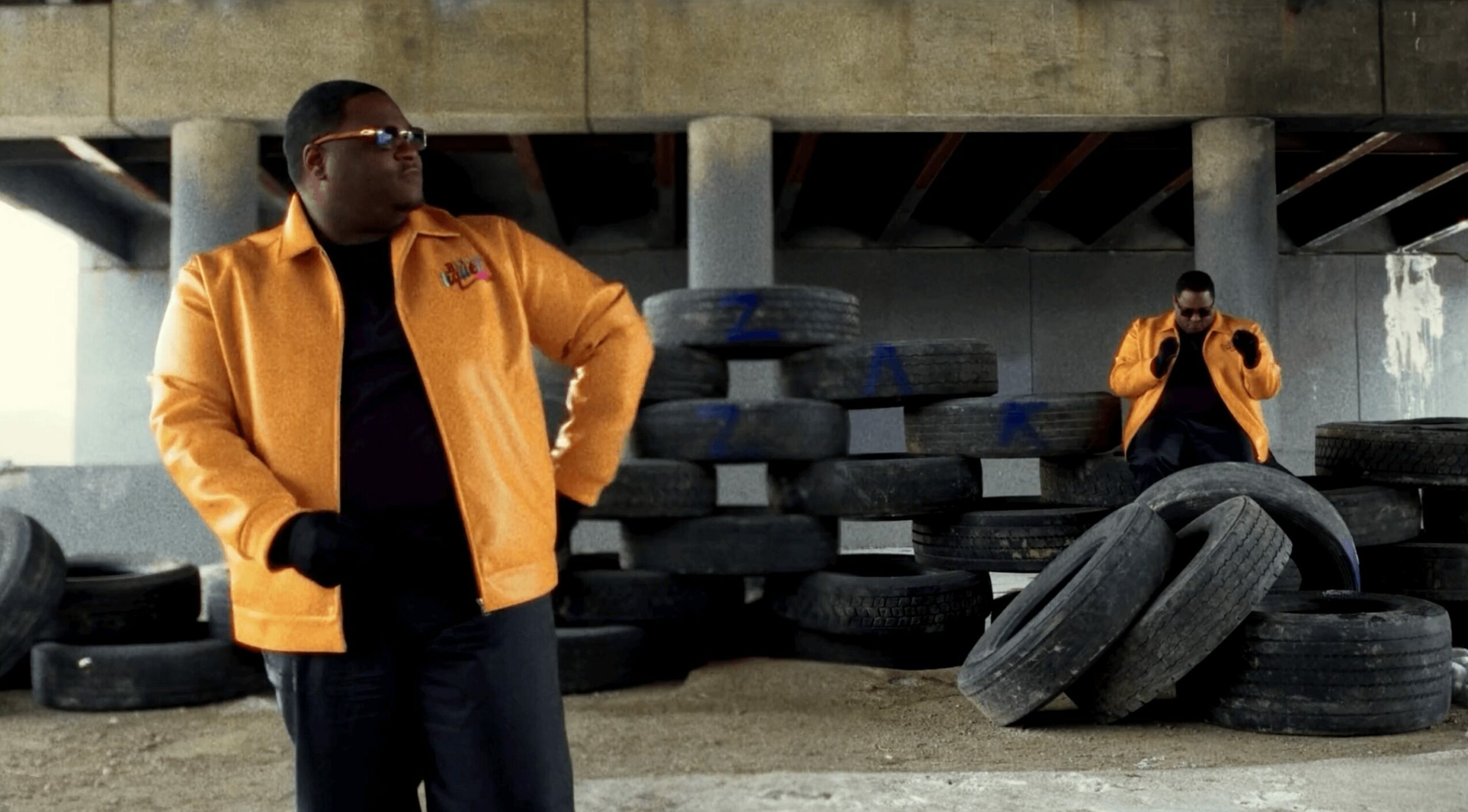 Two men in yellow jackets stand near a pile of tires under a bridge, one looking to the side, the other taking a photo.