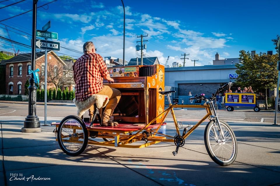 Person playing a piano on a mobile bike platform on a city street with buildings and vehicles in background.