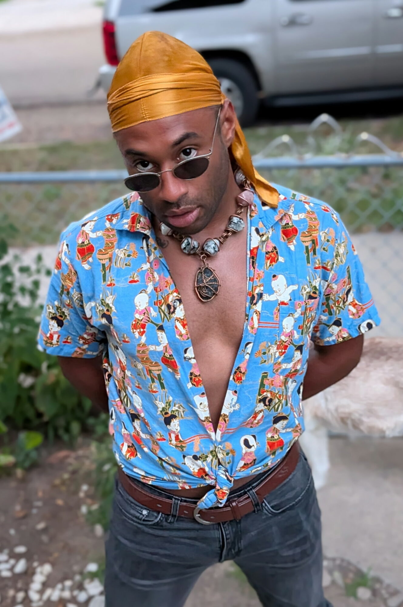 Person wearing sunglasses, a yellow headscarf, a colorful patterned shirt, and jewelry, standing outdoors with a car in background.