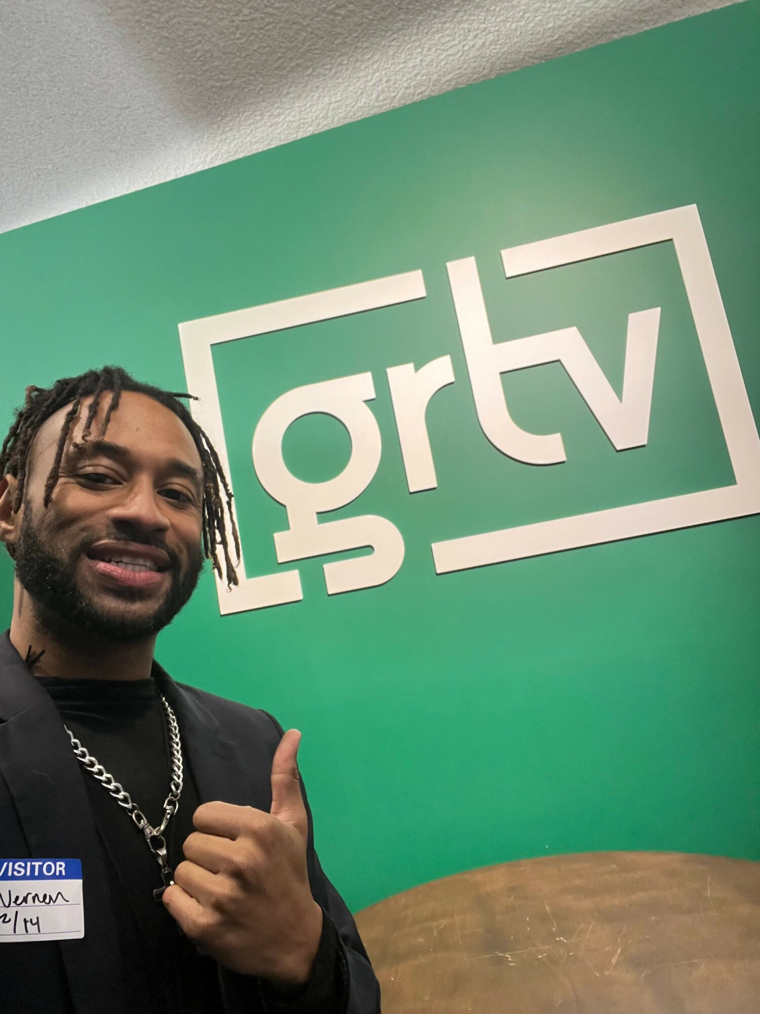 Person with braided hair and beard giving a thumbs-up in front of a green wall with a white logo.
