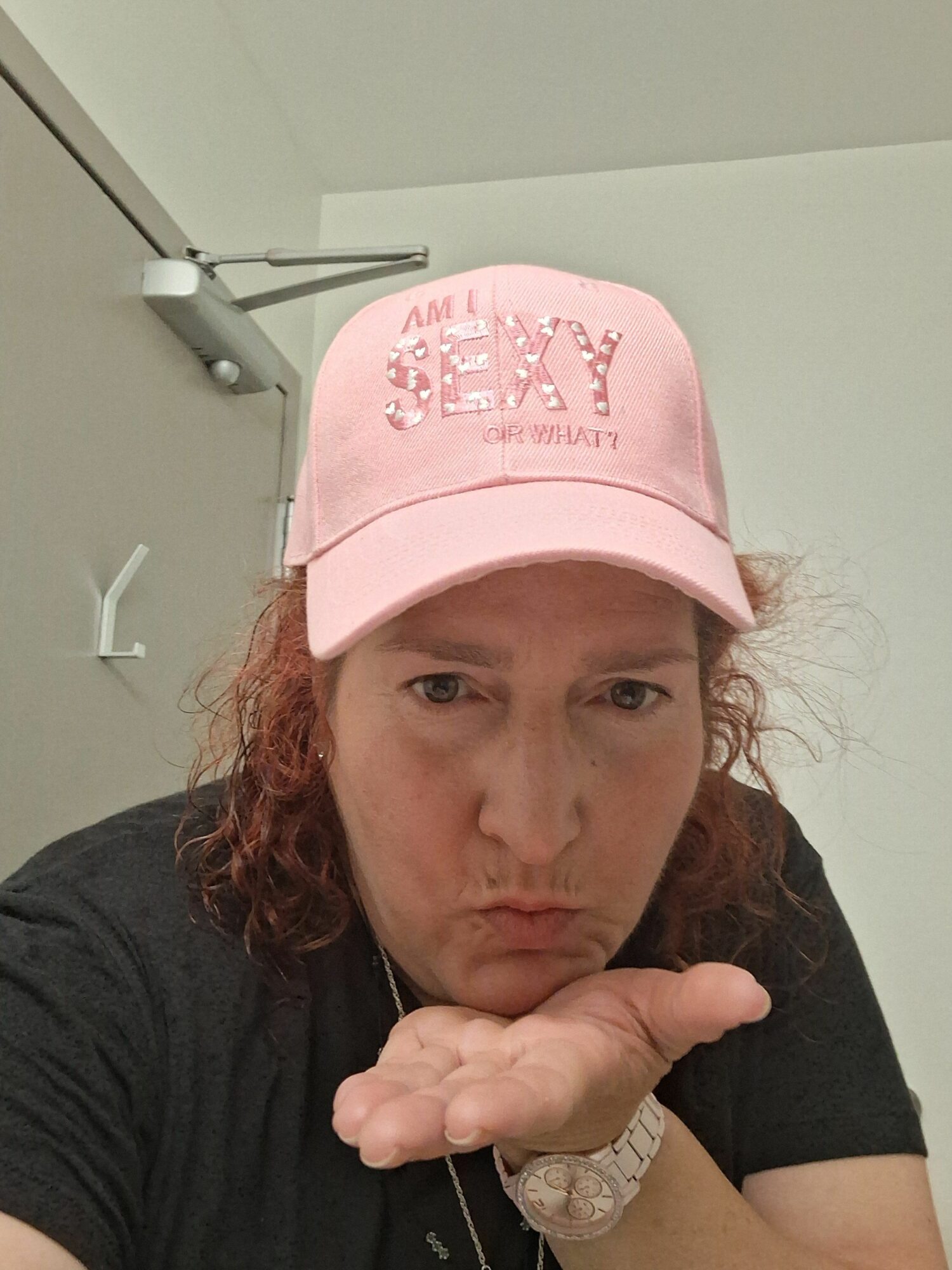 Woman wearing a pink cap with text, resting her head on her hand, in a room with white walls and ceiling light.