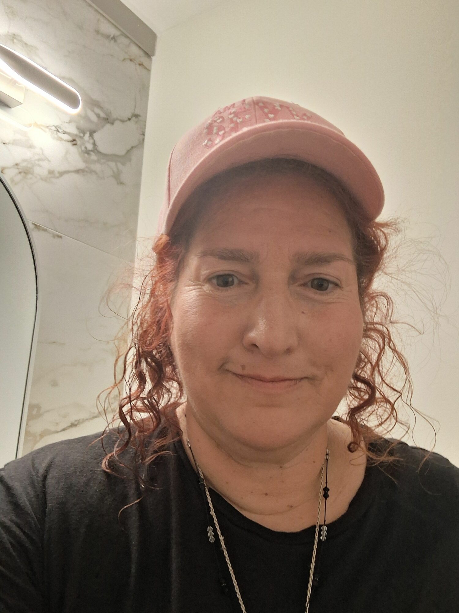 Woman with curly hair wearing a pink cap, black shirt, and necklace, standing indoors with marble wall background.