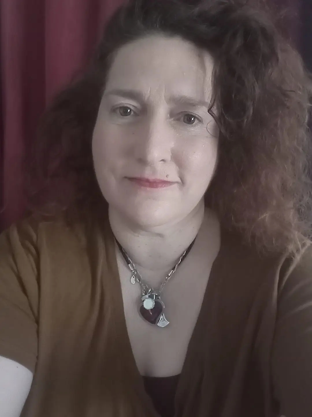 Woman with curly hair wearing a brown top and layered necklaces, looking at the camera with a neutral expression.