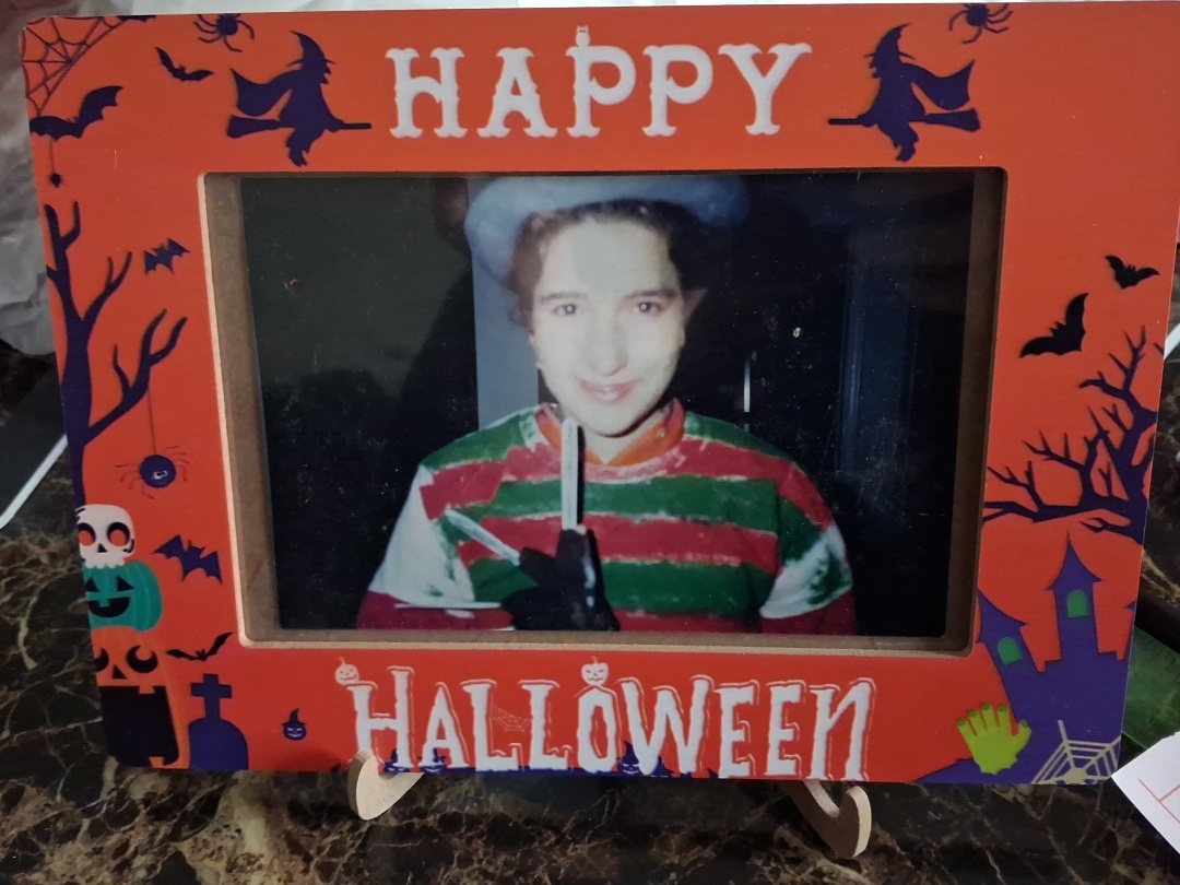 Child in festive sweater holding a knife, smiling, in Halloween-themed frame with bats and trees, orange background.