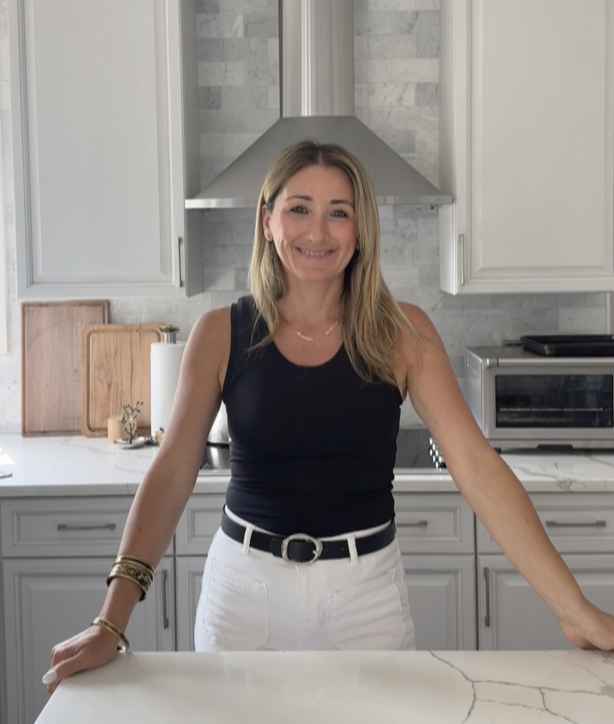 Woman with blonde hair smiling in a kitchen, wearing a black top and white pants, leaning on a white counter.