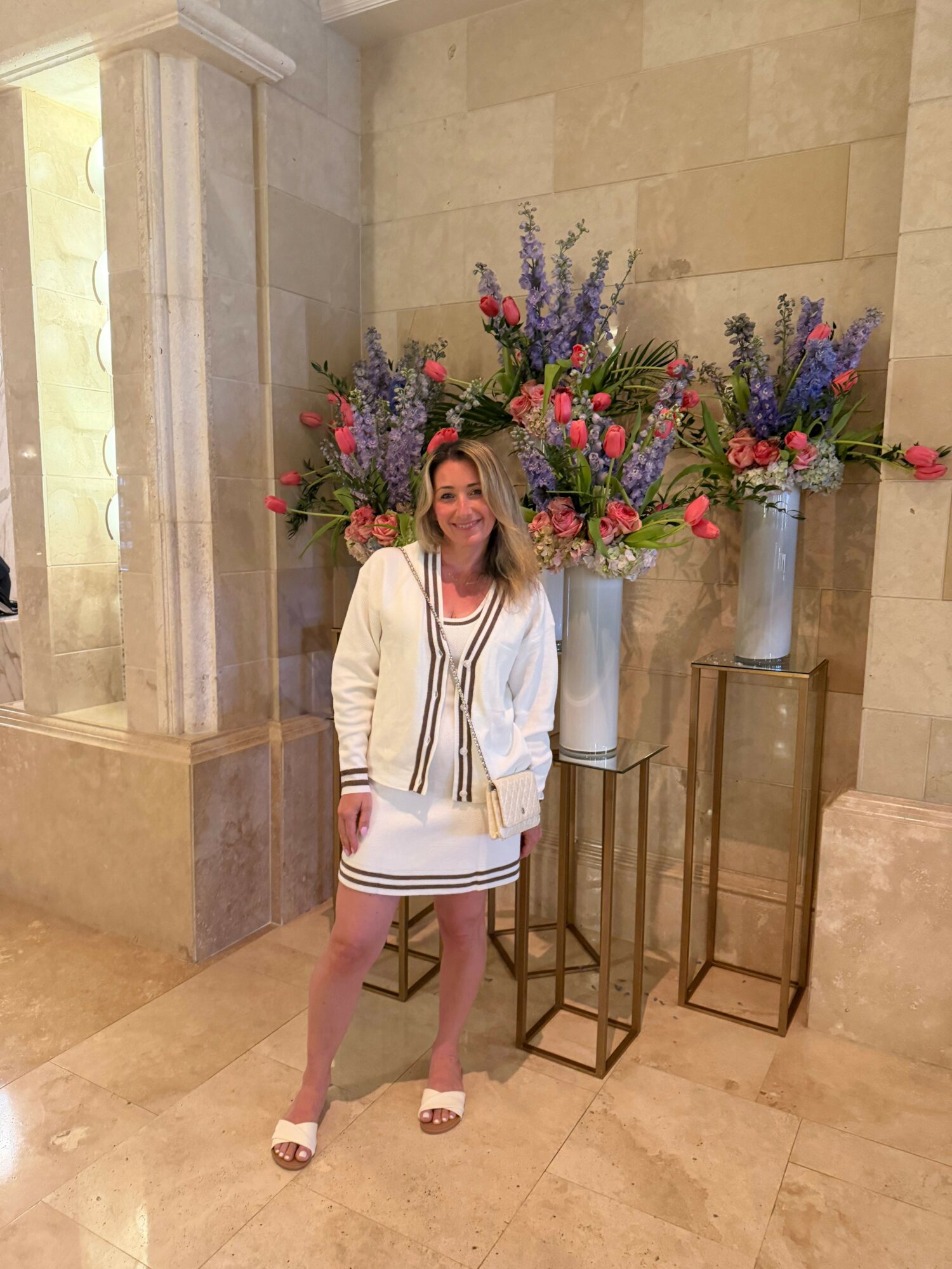 Woman in white dress with black stripes standing near large floral arrangement in a hallway.