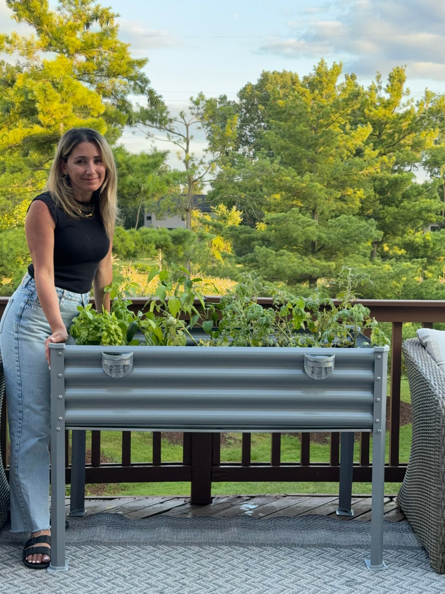 Woman standing on a deck with a large metal planter filled with plants, trees and sky in the background.