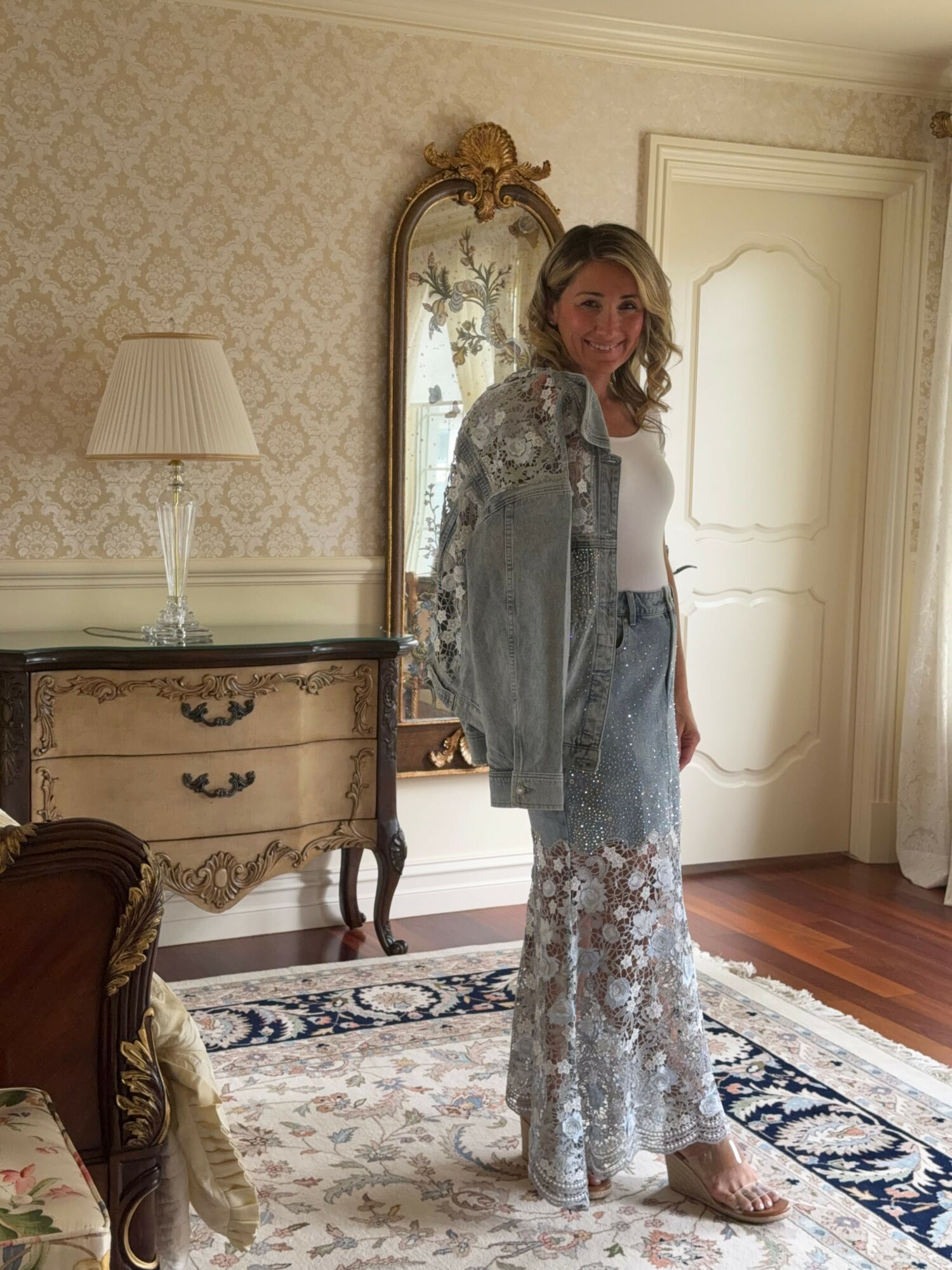 Woman standing in a decorated room with a mirror, dresser, and patterned rug, wearing a long, patterned dress and a light top.