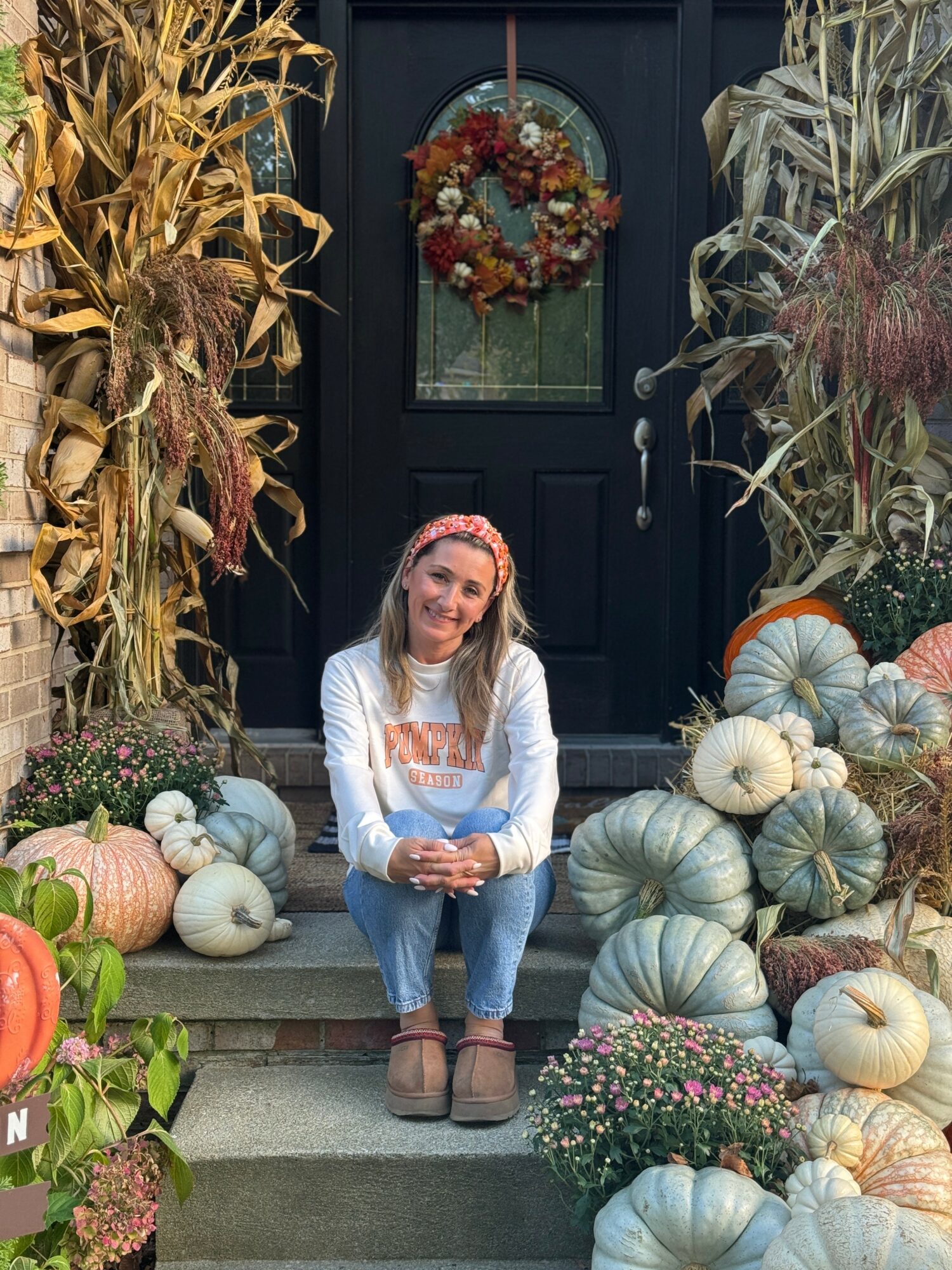 Young woman sitting on steps surrounded by pumpkins and fall decorations, smiling, in front of a dark door with a wreath.