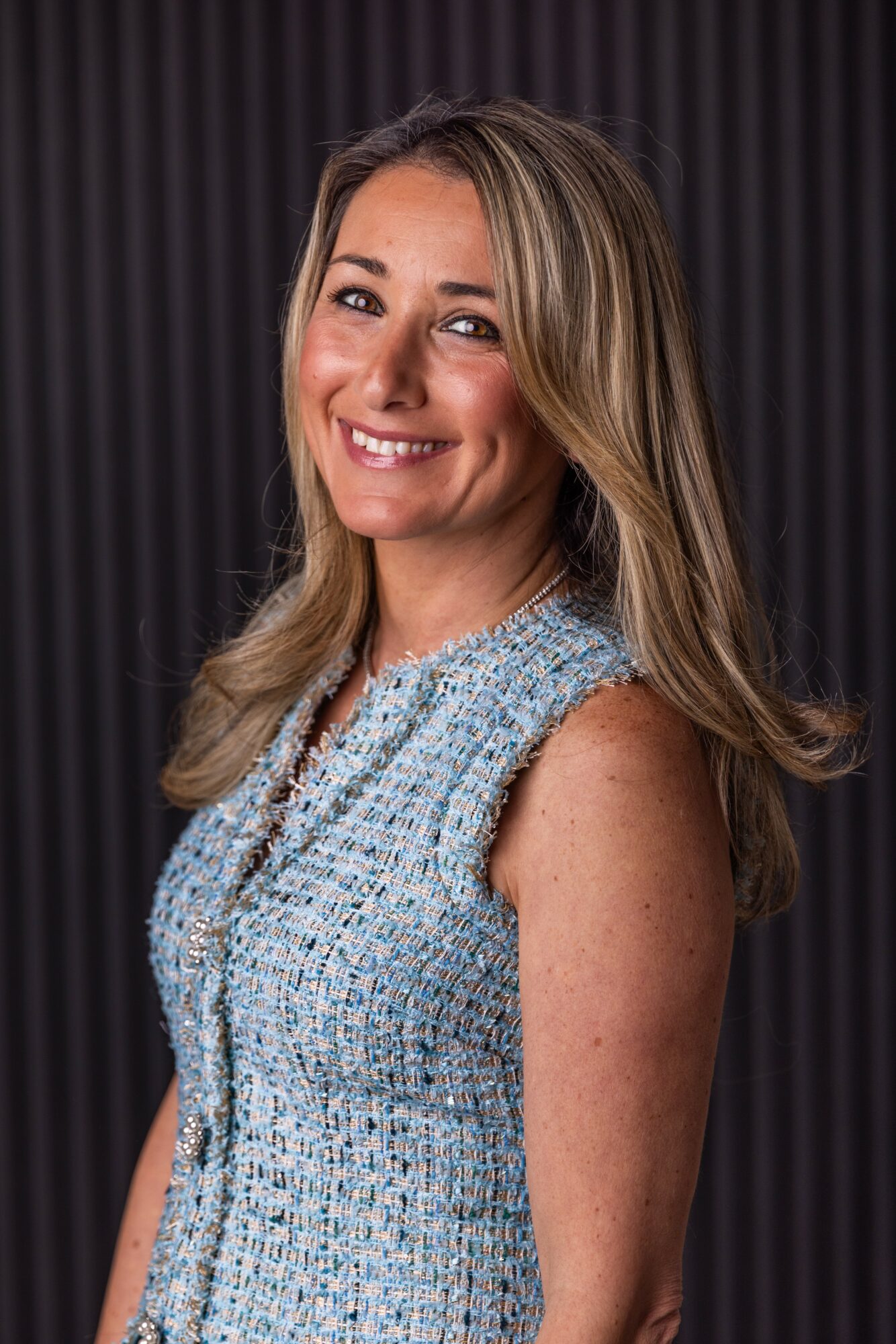 Smiling woman with long blonde hair wearing a sleeveless textured top, standing against a dark background.