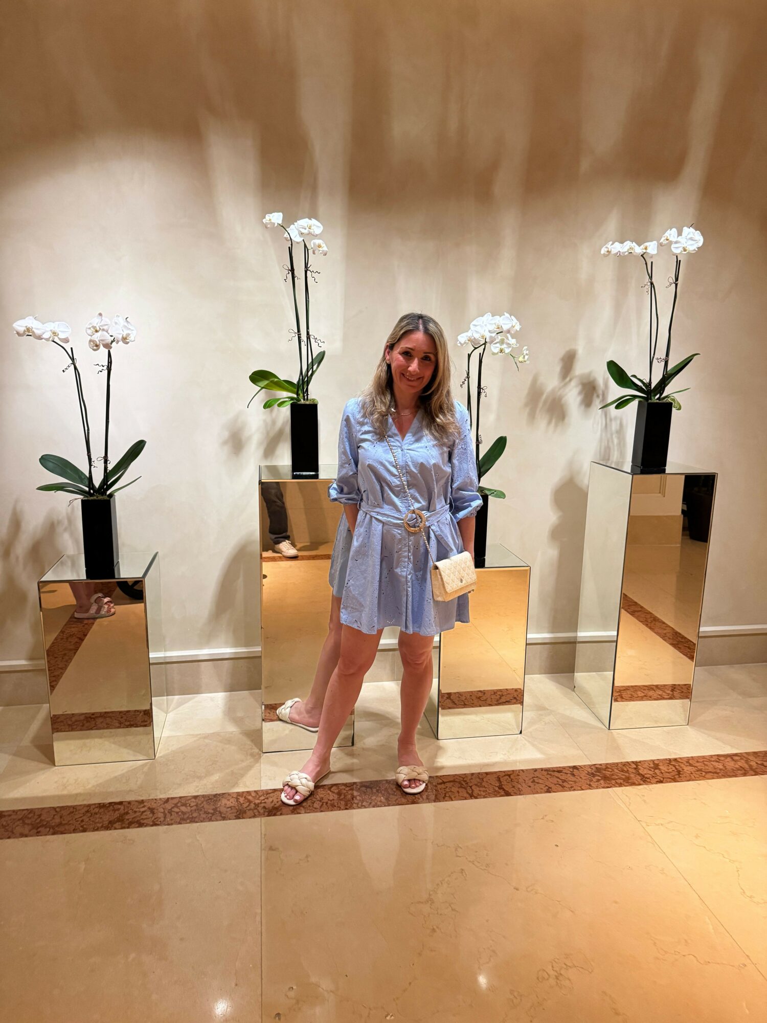 Woman in a light blue dress standing in a decorated interior space with flowers and mirrors.