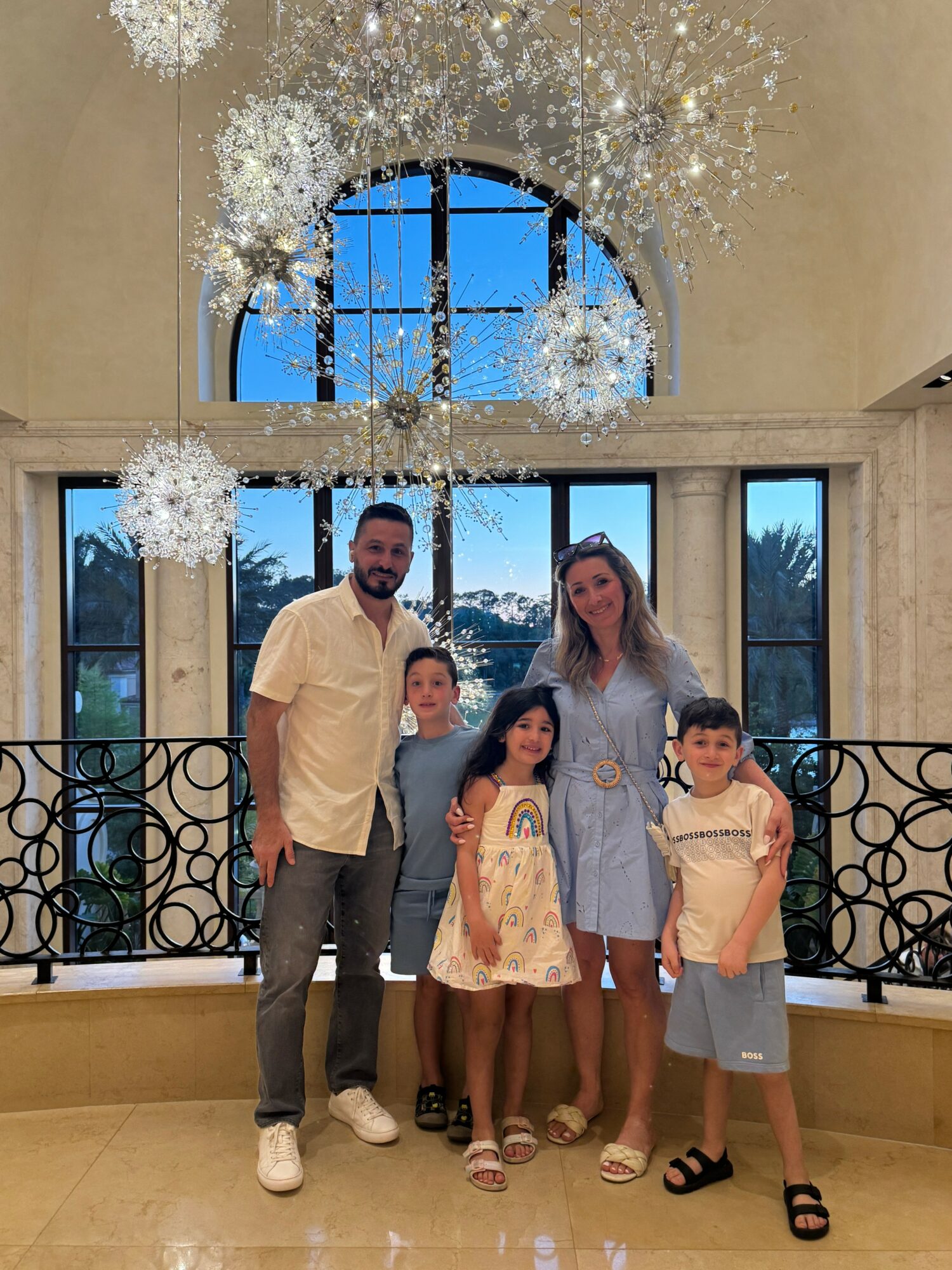Family of five standing indoors with large arched window and decorative ceiling, smiling for photo.