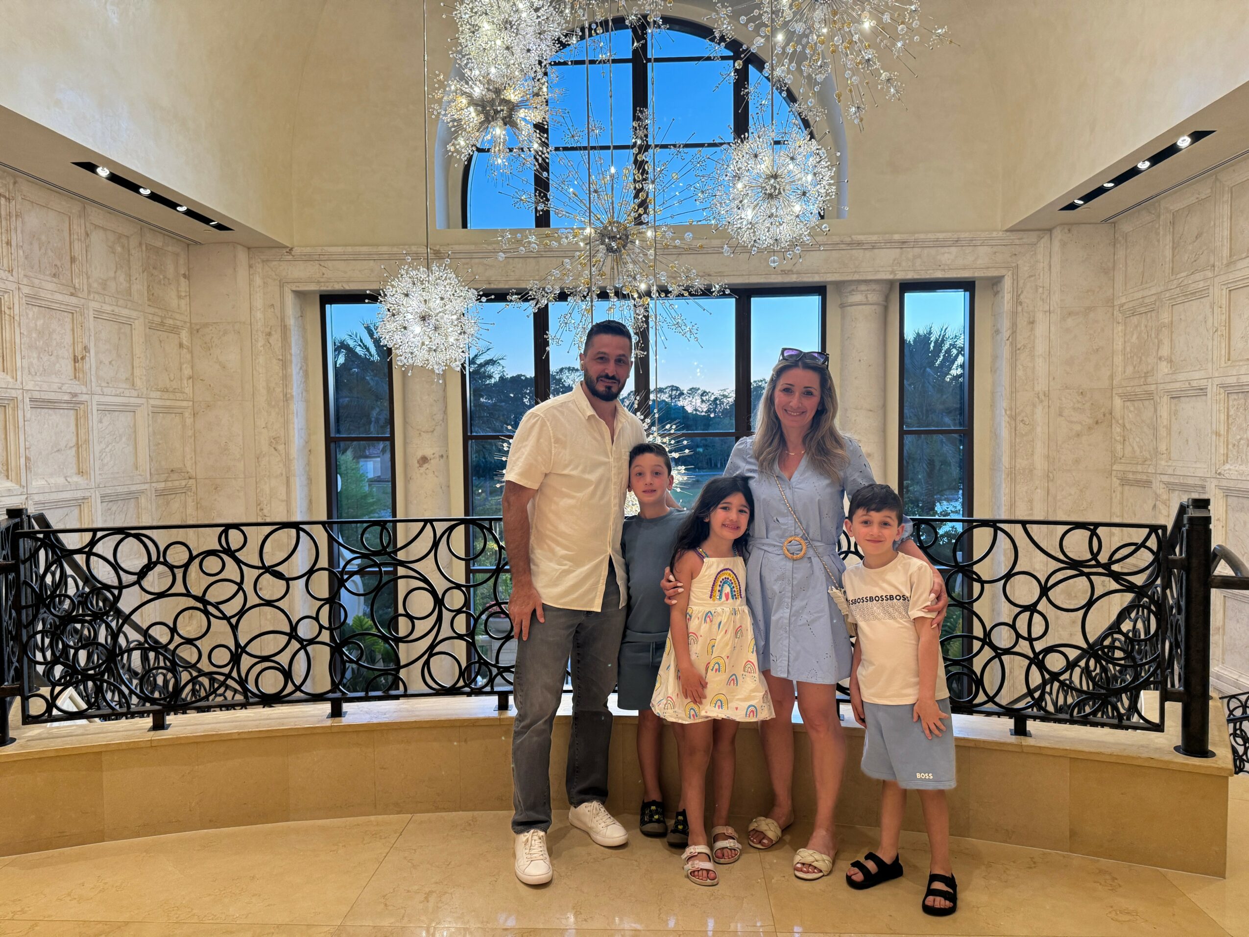 Family of five standing indoors in front of large windows, with decorative hanging lights and ornate railing, smiling at the camera.