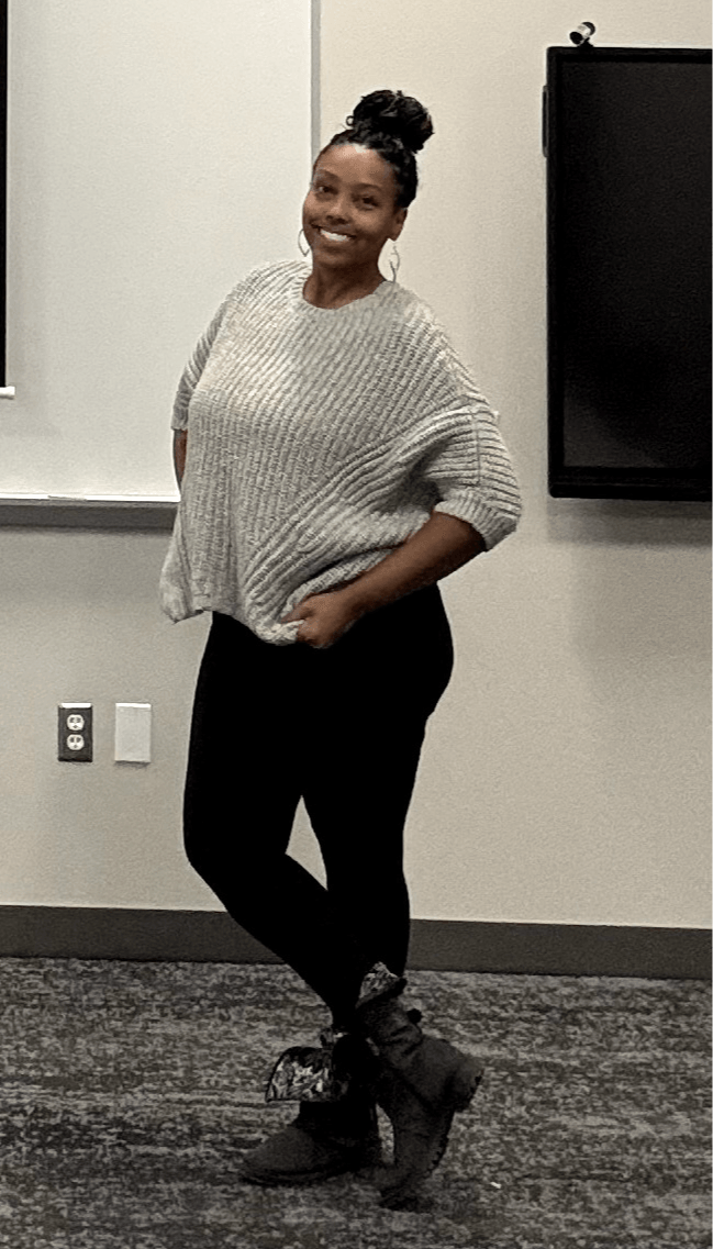 Woman standing in a room, smiling, wearing a loose sweater, black pants, and boots, with a black screen behind her.