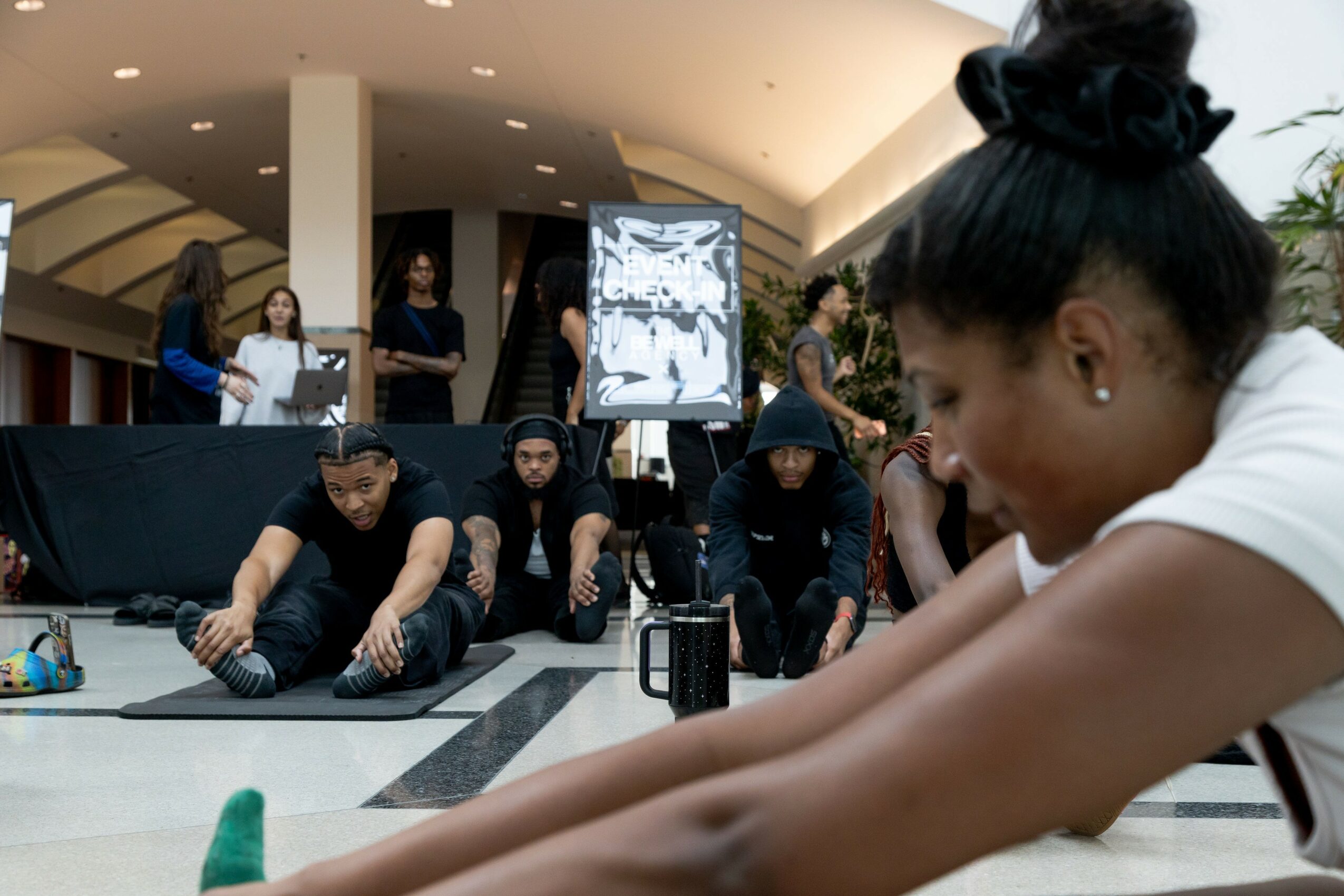 A woman stretches on the floor while a group of people sit behind her, with a display screen in the background.