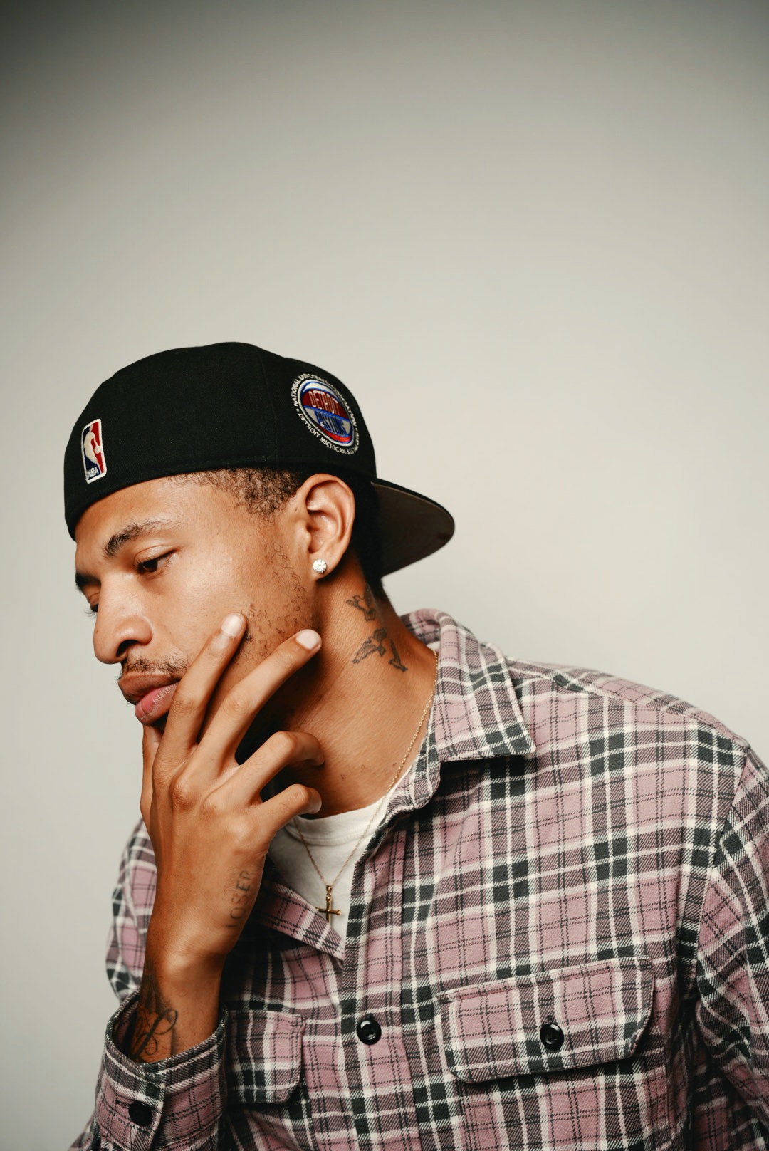 Young man wearing a black cap with NBA logo, plaid shirt, and a white t-shirt, touching his face thoughtfully.