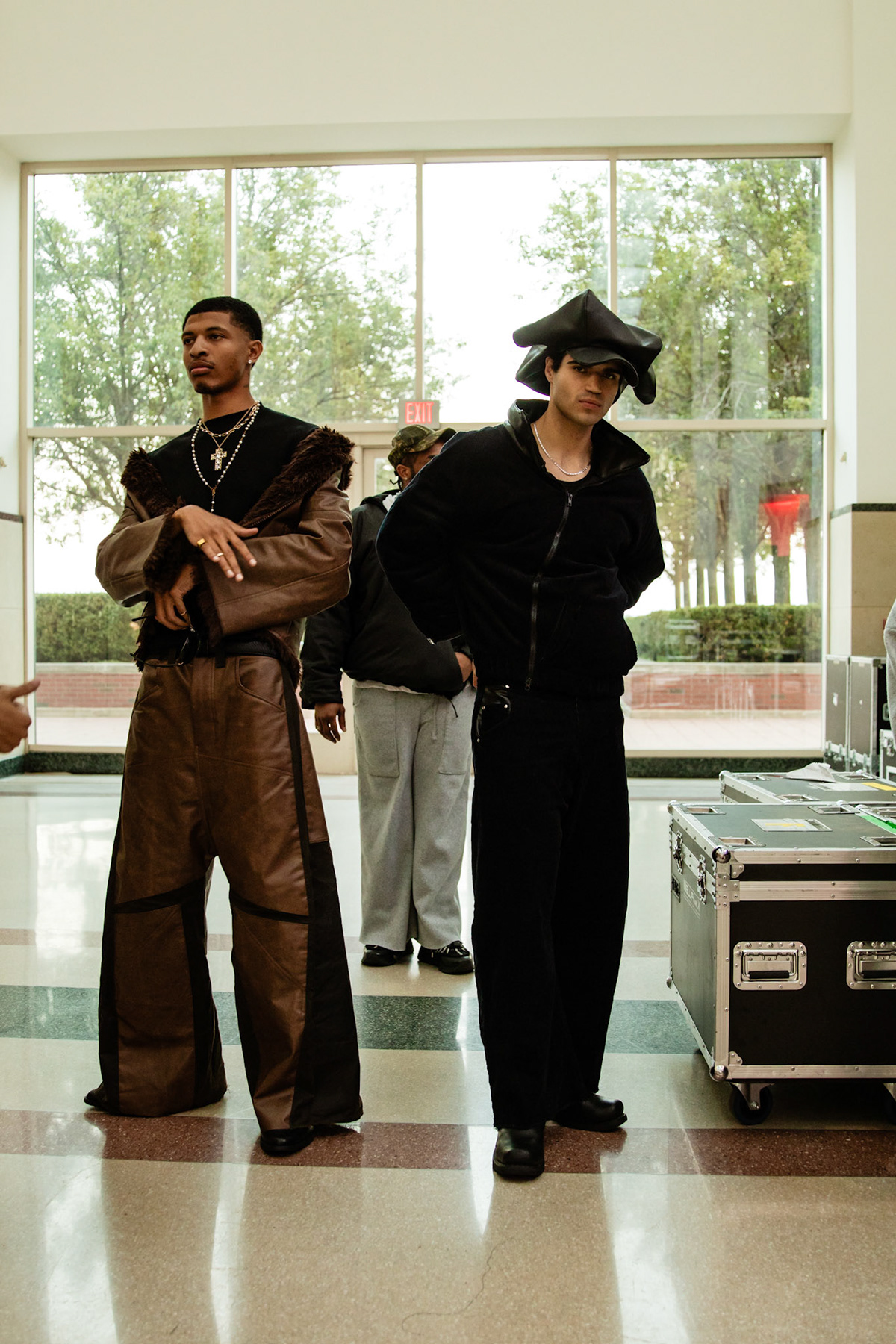 Two people standing indoors in front of large window, one with arms crossed, the other wearing a hat and dark clothing.