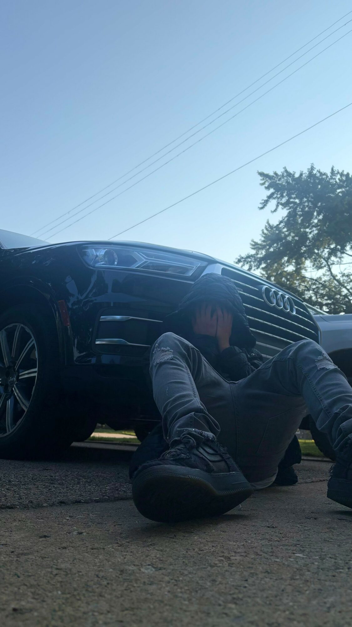 Person sitting on ground with legs extended, leaning against a large black vehicle, outdoors with trees and power lines in background.