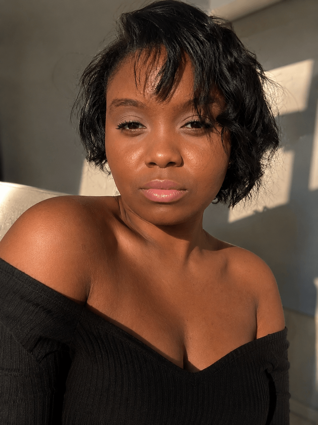 Young woman with short, wavy black hair, wearing a black off-shoulder top, looking at the camera with sunlight on her face.