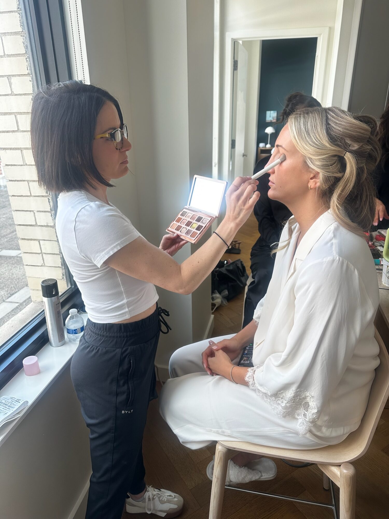 Makeup artist applying makeup to a seated woman with blonde hair in a white outfit, near a window with natural light.