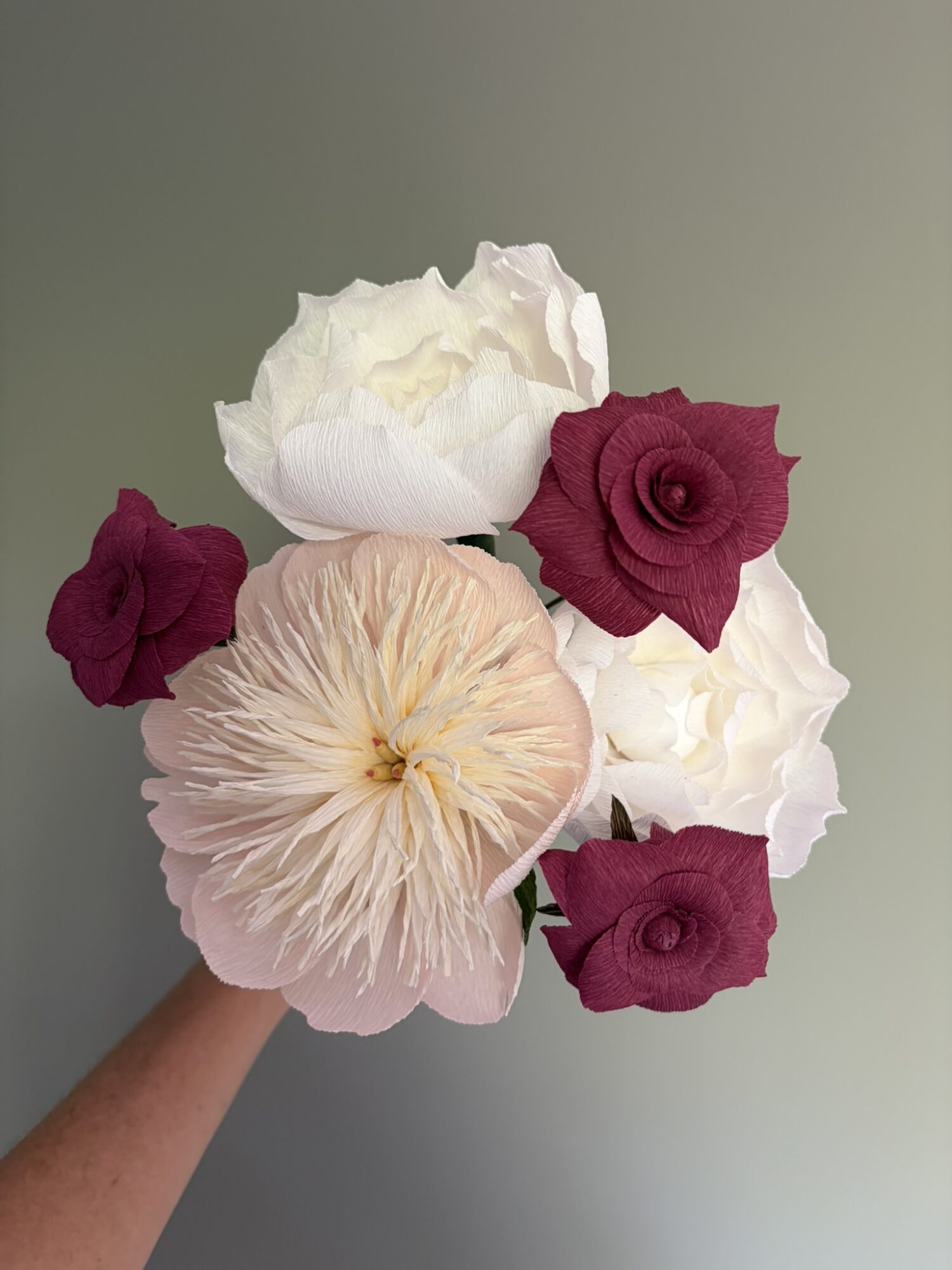 Person holds a bouquet of white, pink, and dark red flowers against a plain background.