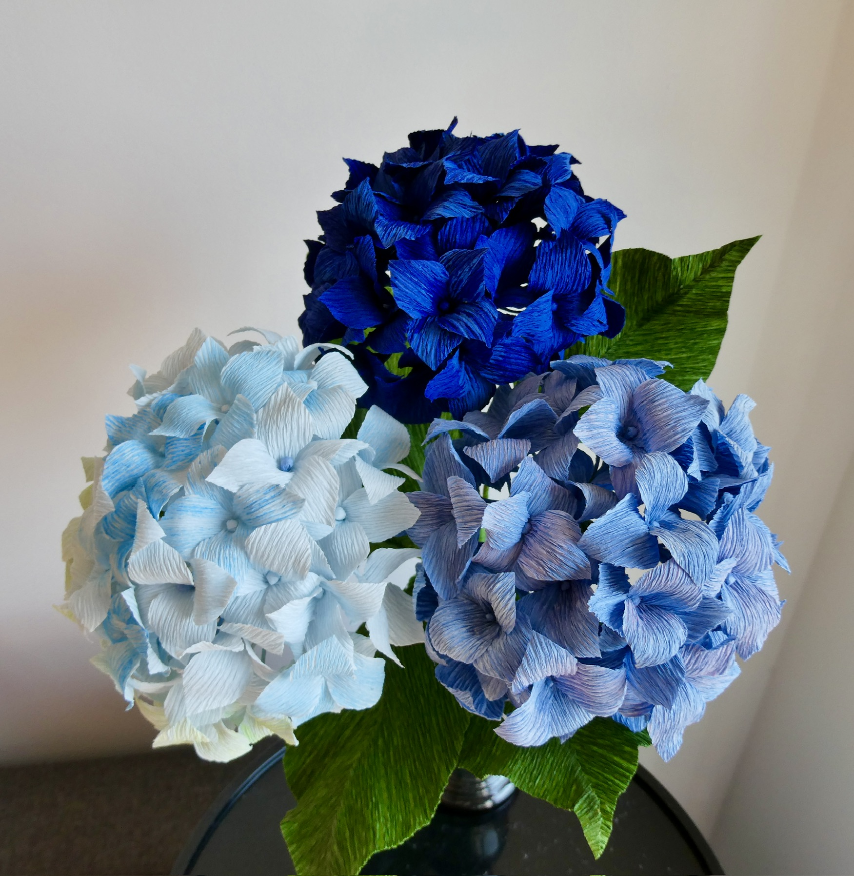 Three blue and white hydrangea flowers with green leaves in a black vase, against a plain background.