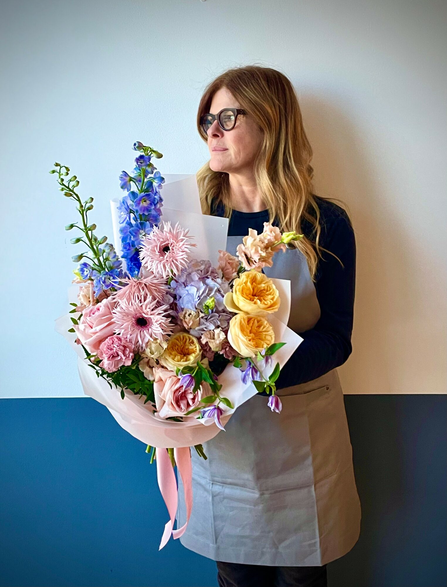 Woman with glasses holding a large bouquet of pink, yellow, and purple flowers, standing against a plain wall.