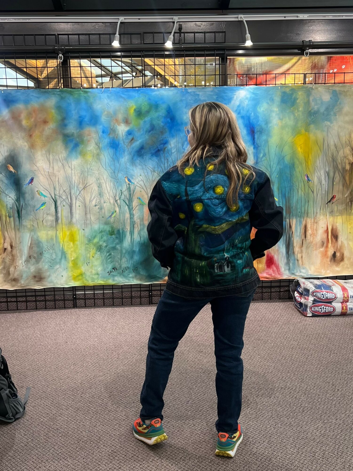 Person with long hair wearing a dark jacket and jeans stands in front of a colorful abstract mural.