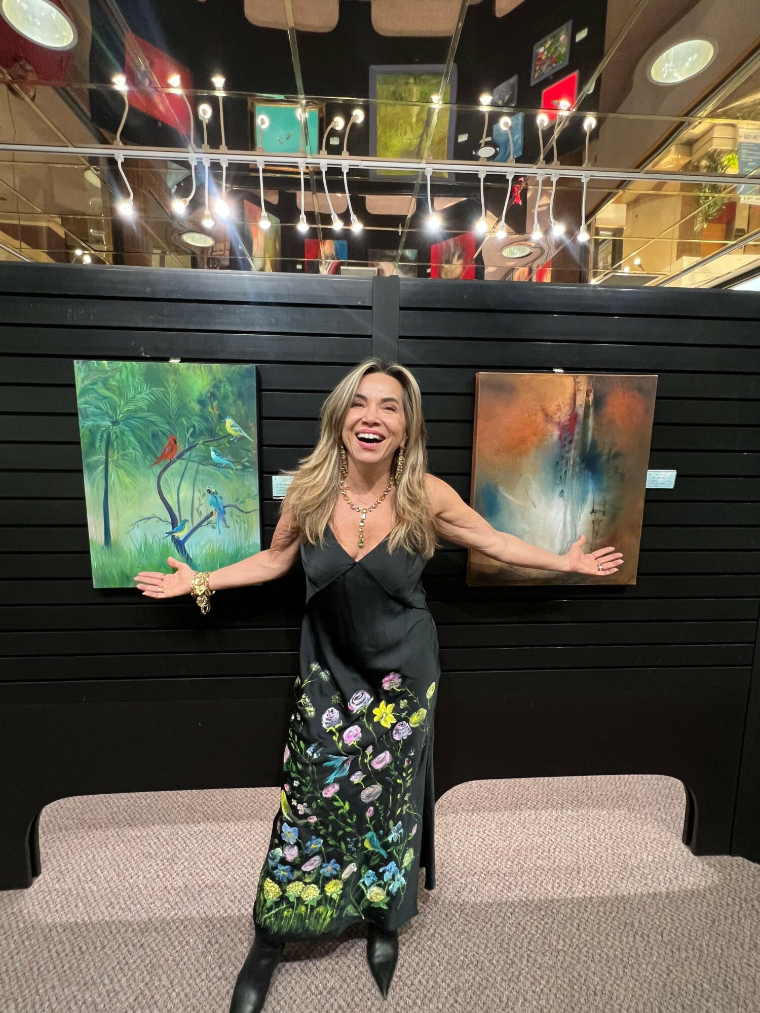 Woman with long blonde hair smiling and waving, standing in front of colorful paintings, in an indoor setting.
