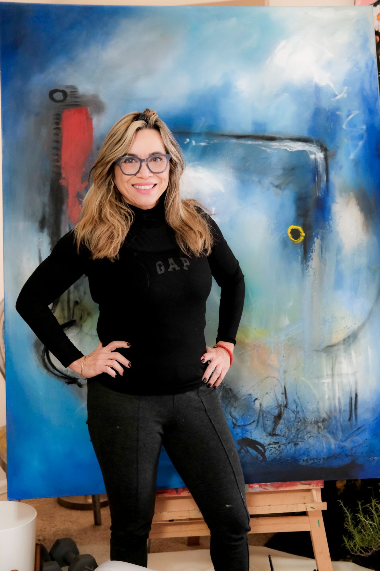 Young woman with glasses and wavy hair smiling, standing in front of a colorful abstract painting, hands on hips.