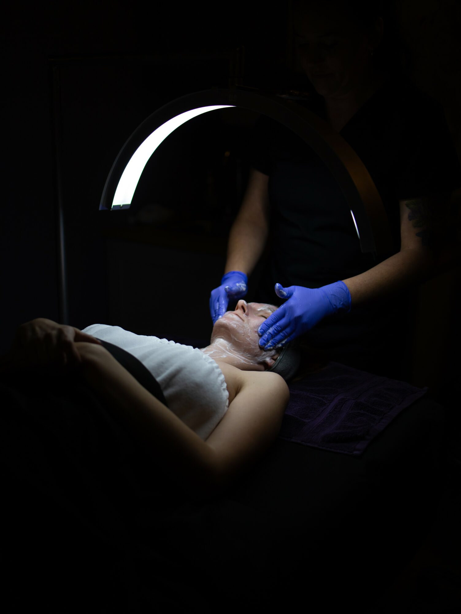 Person lying down with head supported, undergoing a medical procedure with a healthcare professional wearing blue gloves, in a dark room.