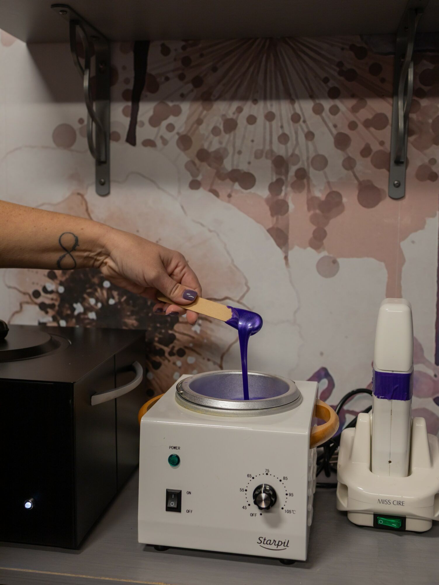 Person's hand holding a purple object over a white device with a dial, on a table with a wall and shelf in background.