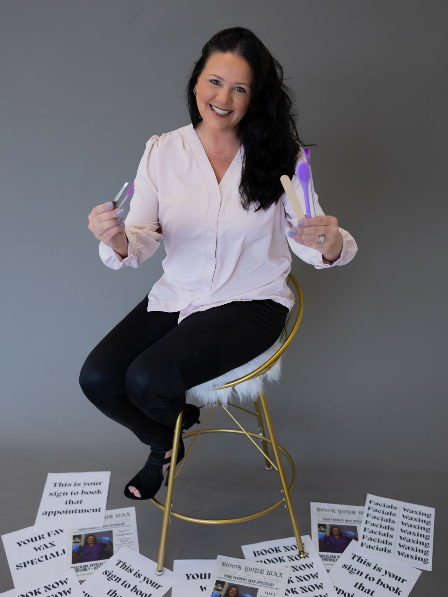 Woman with long dark hair smiling, sitting on a gold chair, holding two purple objects, against a gray background.