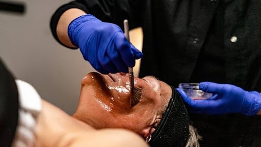 Person receiving a cosmetic or medical procedure on their face, with practitioner wearing blue gloves, holding tools near face.