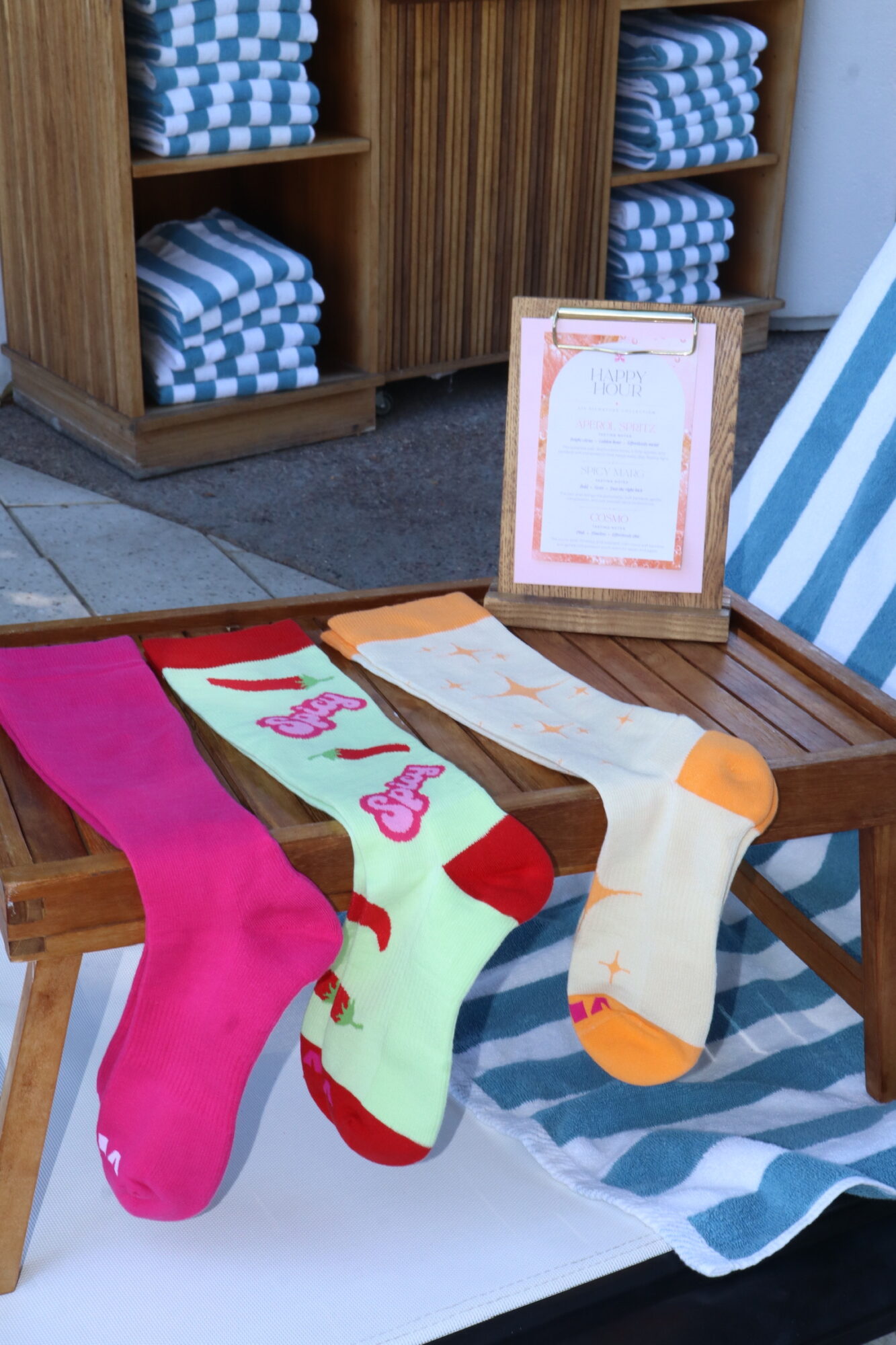 Four colorful socks are displayed on a wooden bench outdoors, with a framed sign and shelves of folded towels in the background.