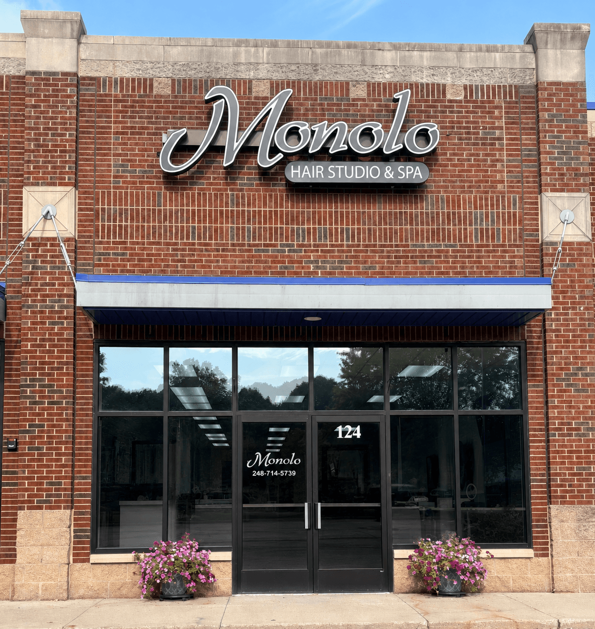 storefront with sign 'Monolo Hair Studio & Spa', glass door, two flower pots, brick building, blue awning, street view