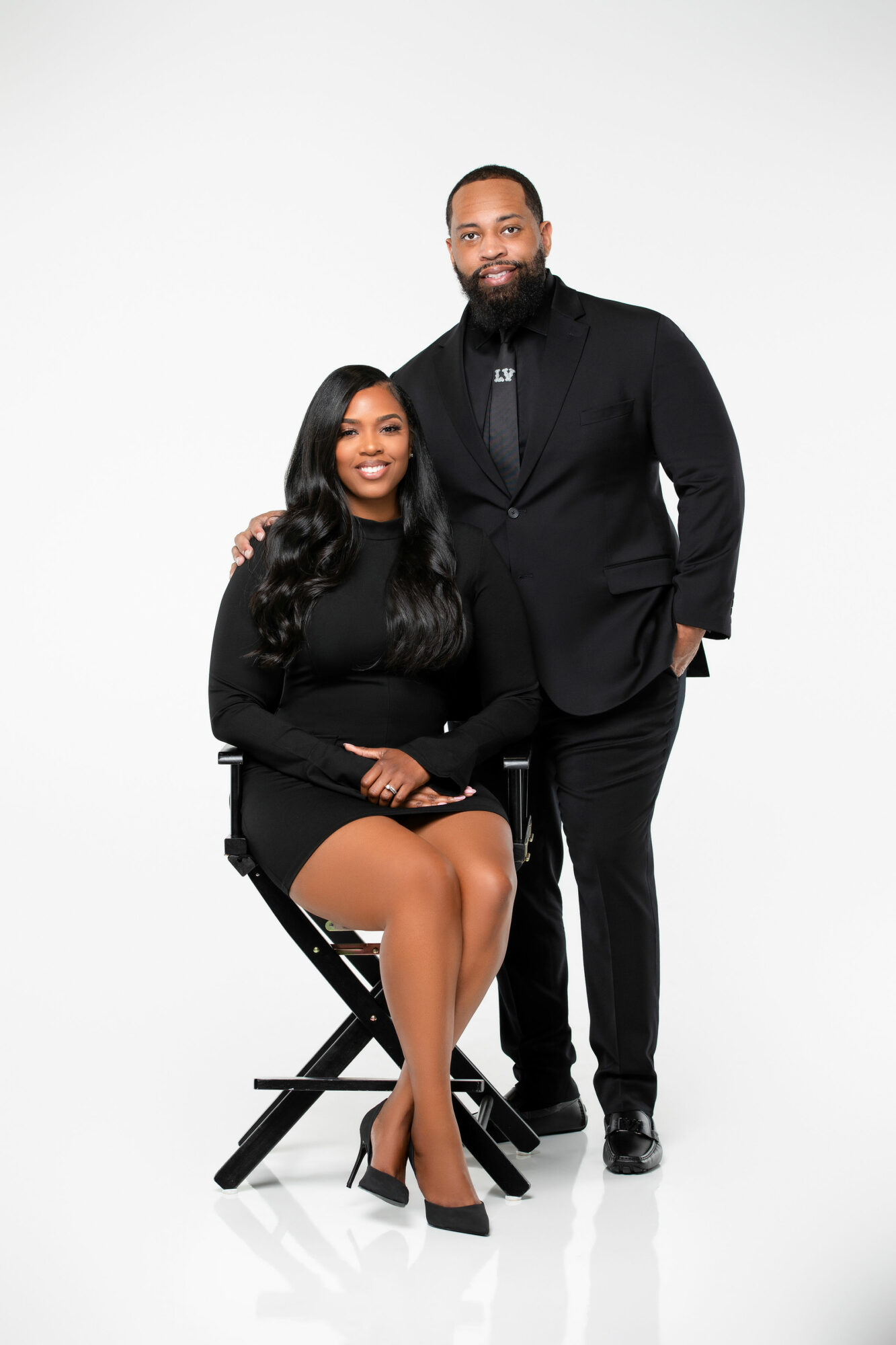 A man and woman pose together, the woman seated on a stool and the man standing behind her, both smiling.