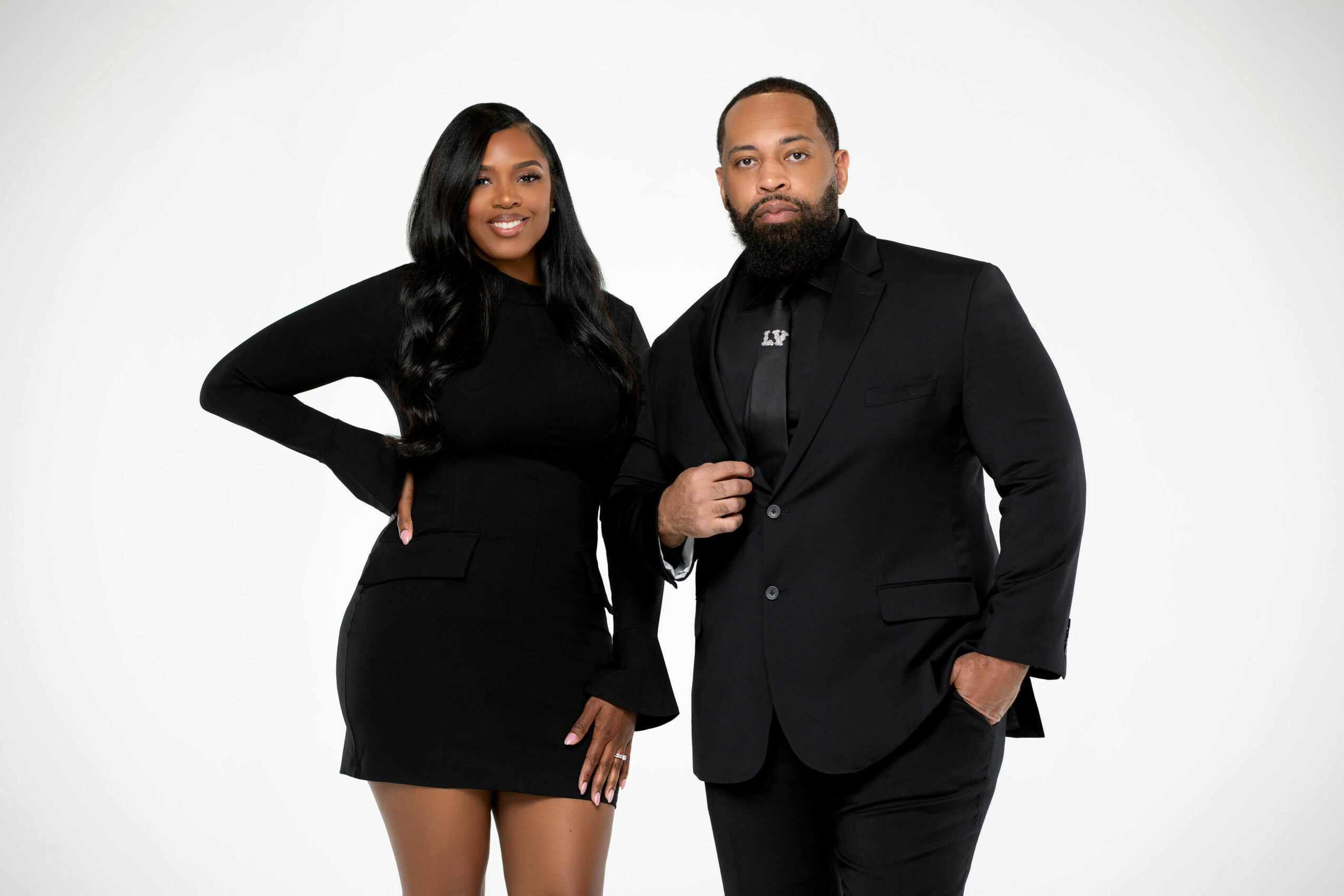 Two people in black formal attire standing against a white background, smiling at the camera.
