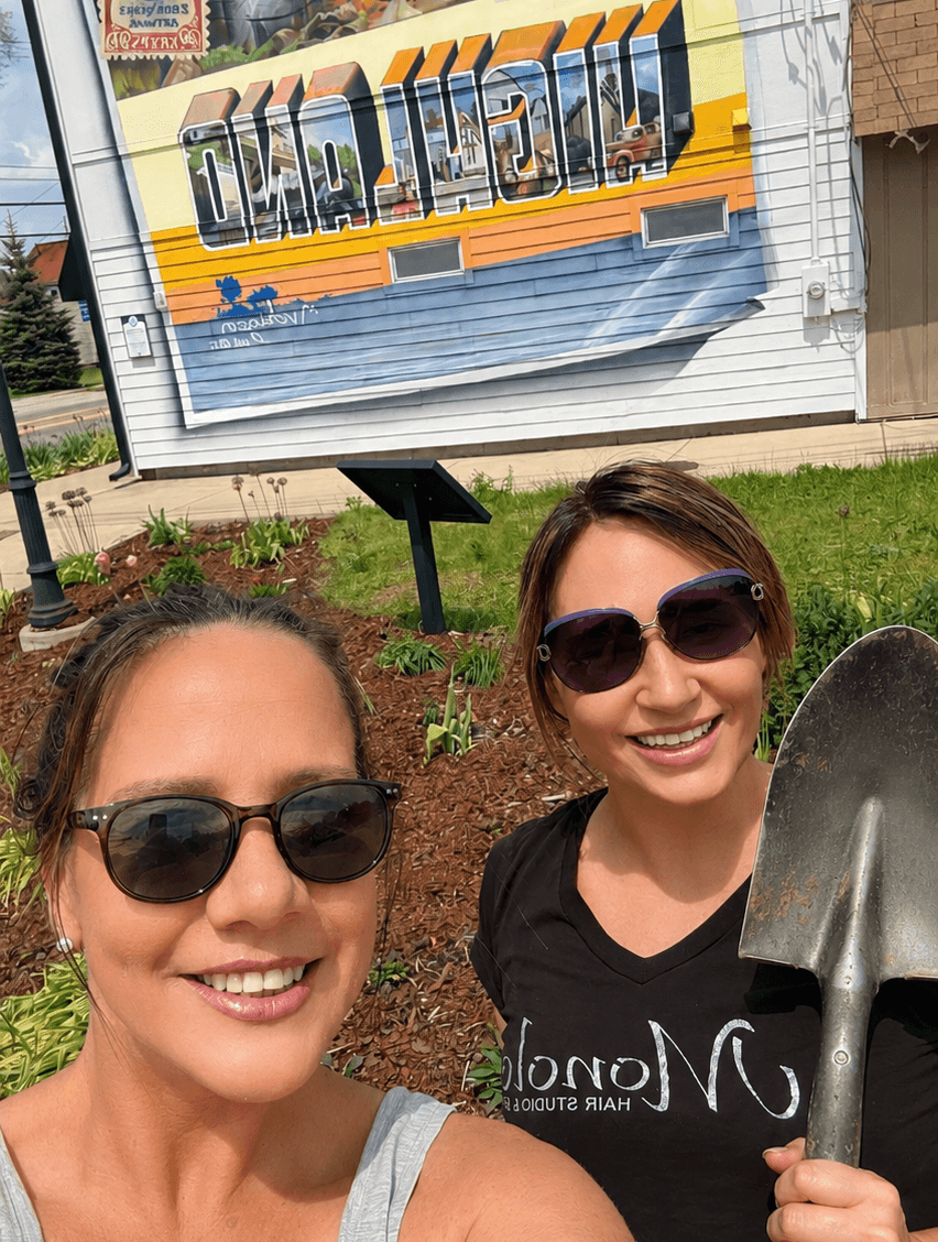 Two women wearing sunglasses smiling outdoors with a mural and garden in the background.