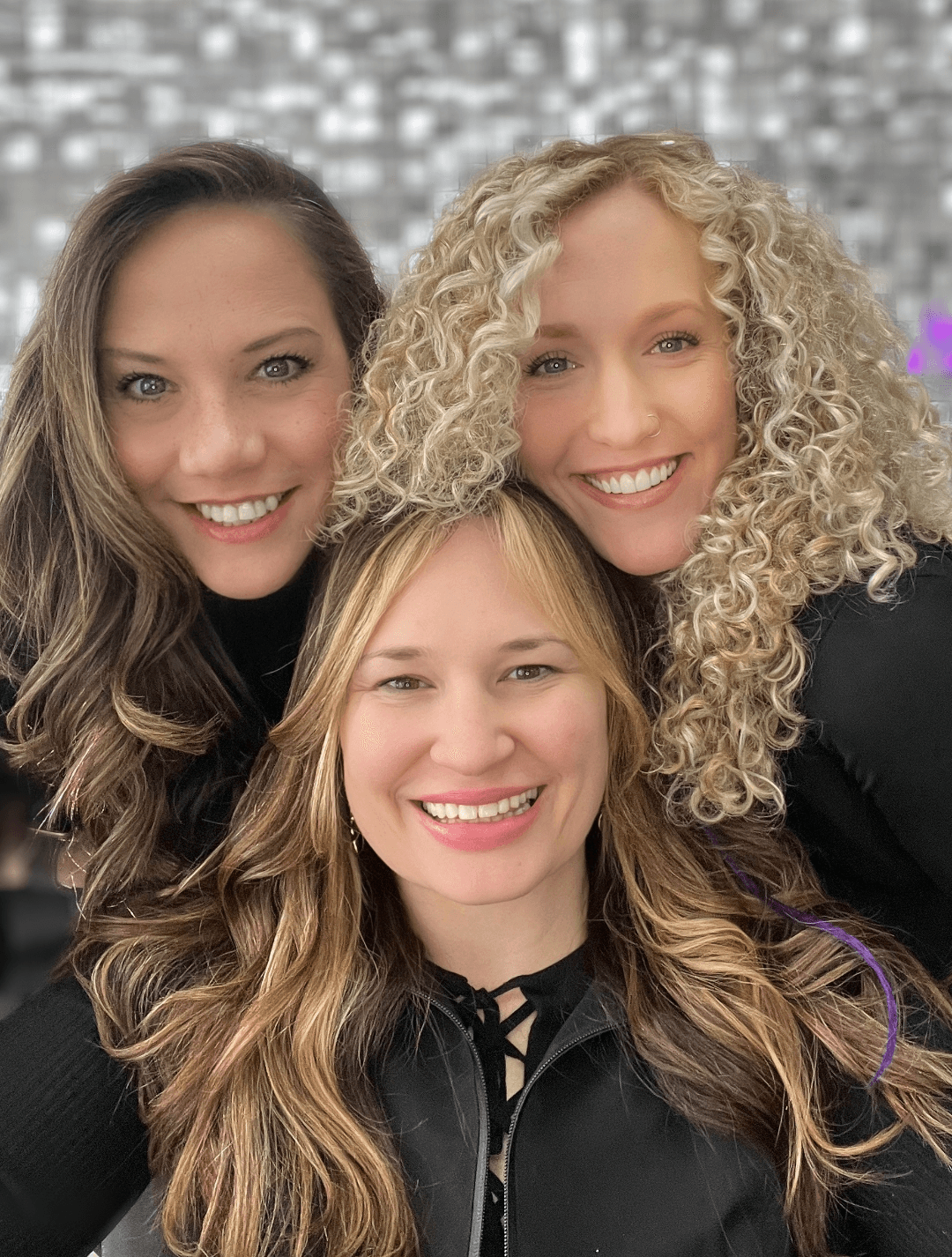 Three women smiling closely together, with a blurred background of lights and decorations.