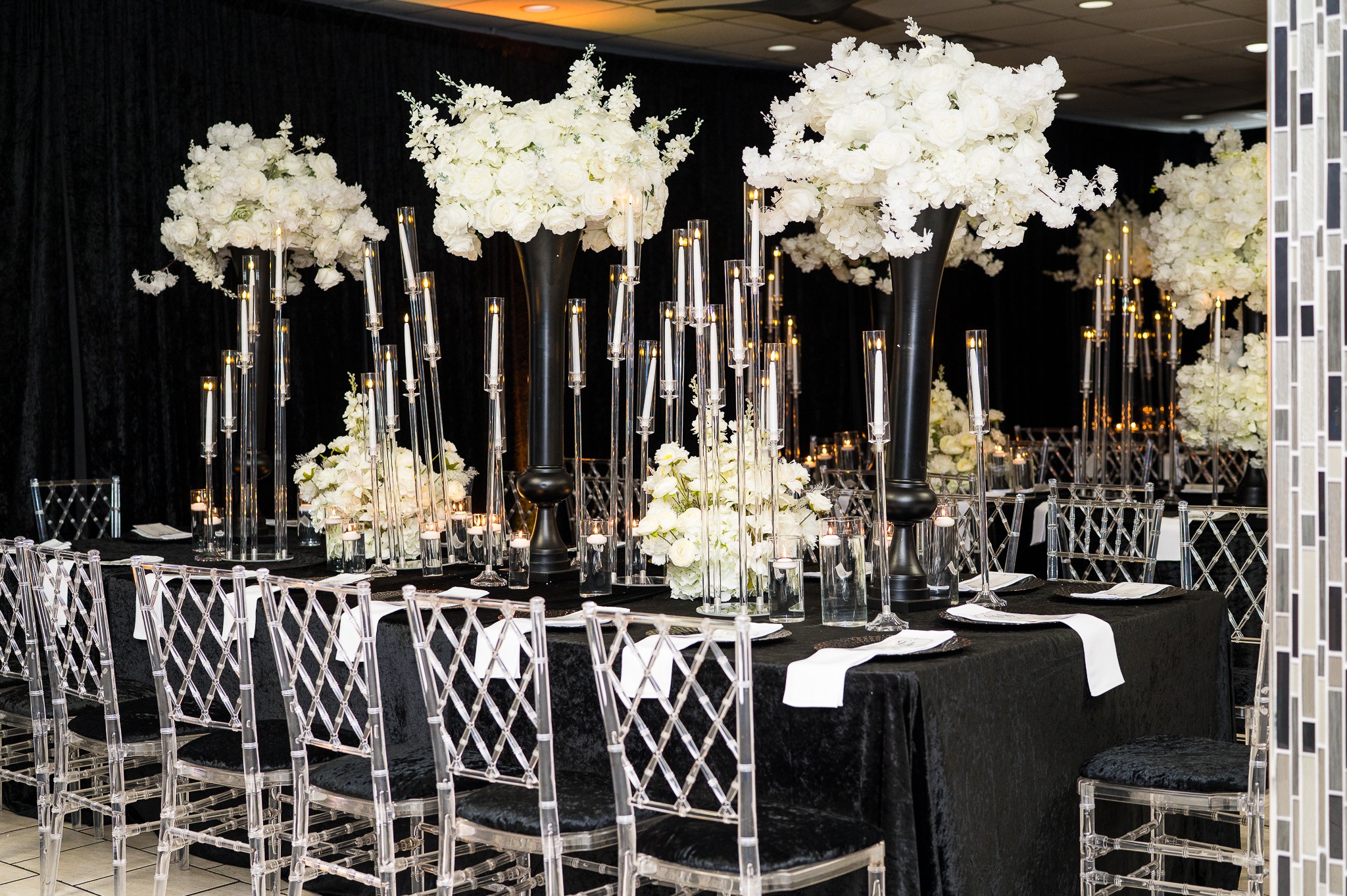Elegant table setting with tall floral centerpieces and black tablecloths, silver chairs, and a dark background.
