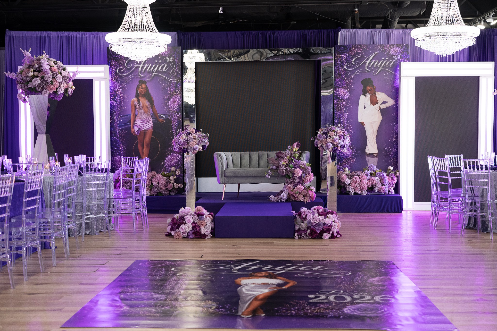Event stage with purple and pink floral decorations, two large posters of a woman, and a central sofa, in a decorated hall.
