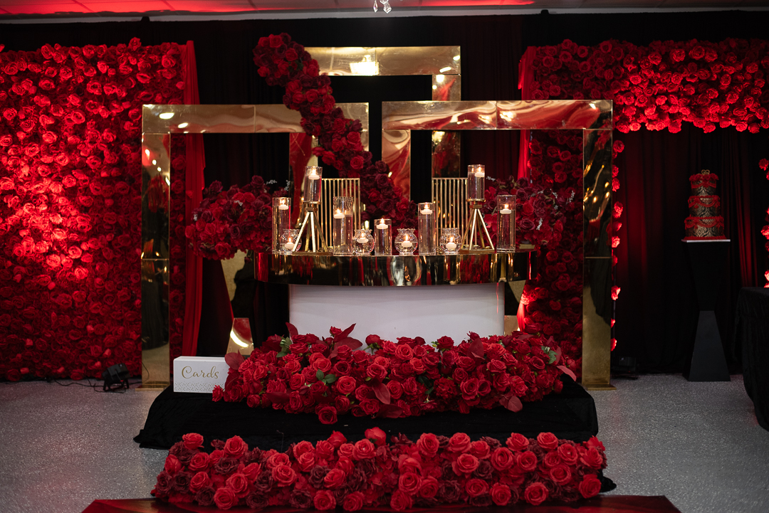 Decorative setup with red flowers, gold frames, and glassware on a table, with a red floral backdrop.