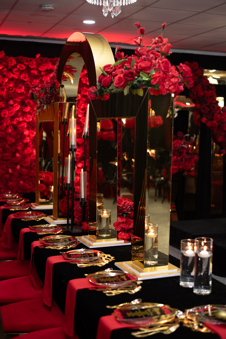 Elegant table setting with black and red tablecloths, gold accents, tall floral centerpieces, and glassware, in a decorated event space.