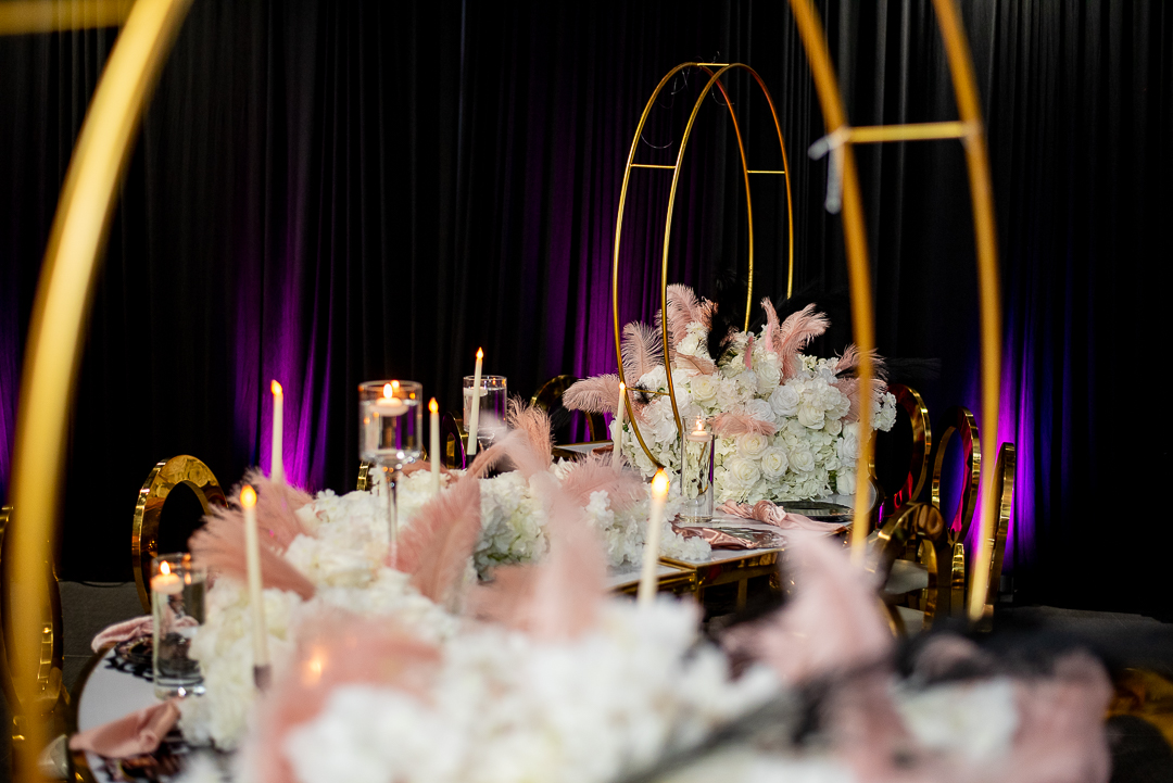 Elegant table setup with floral arrangements, candles, and gold decorative arches, illuminated by purple lighting, against a black curtain backdrop.