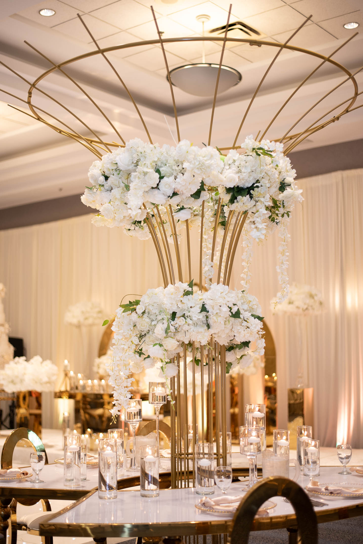 Elegant table centerpiece with white flowers and gold accents, set in a decorated event space with warm lighting.